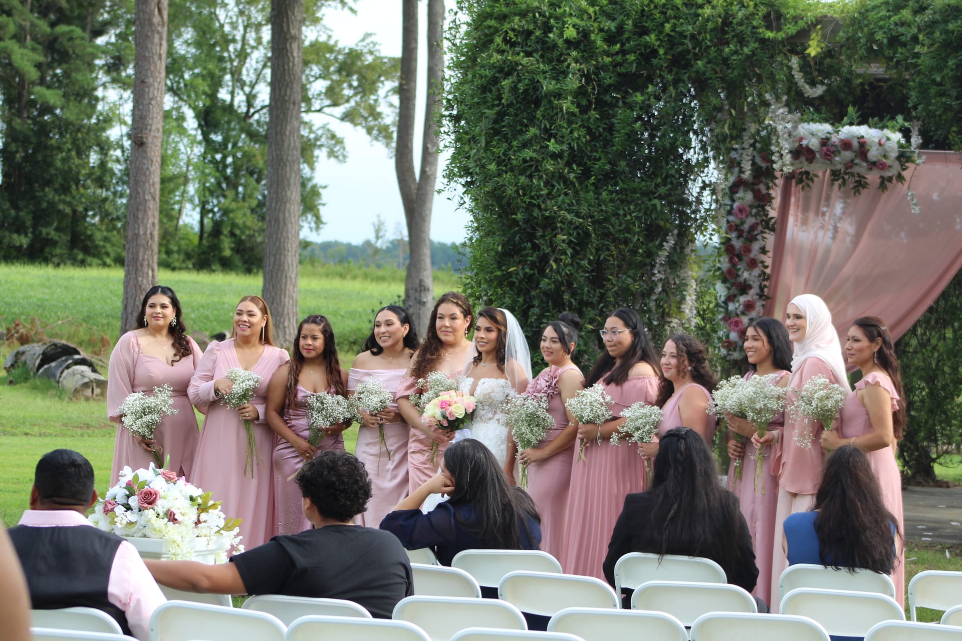 Bride in white gown and veil stands with eleven bridesmaids in dusty-rose dresses holding baby's breath bouquets on the ceremony lawn in front of a floral-draped arch at Scarborough House.