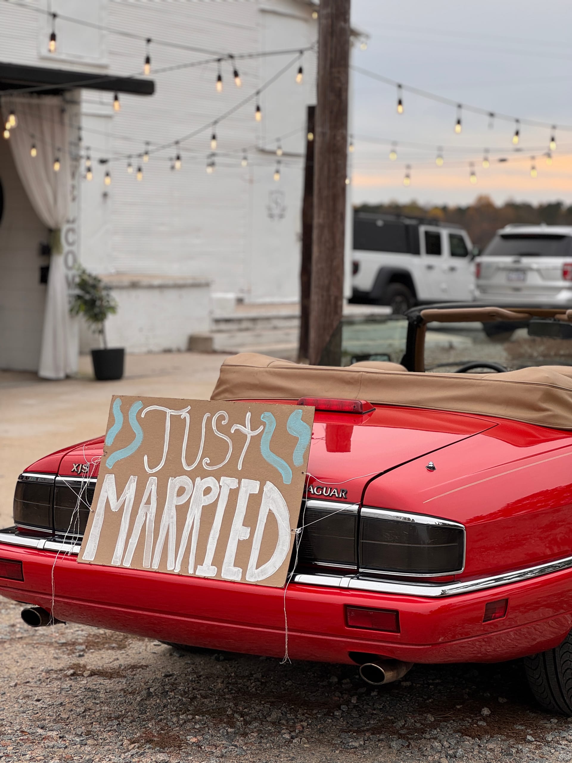 A classic red Jaguar convertible adorned with a handmade Just Married cardboard sign parked in the courtyard of Scarborough House wedding venue, framed by warm string lights at dusk.