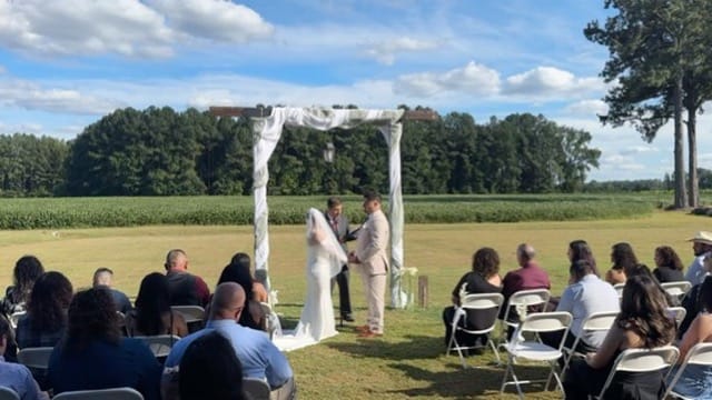 A bride and groom exchange vows beneath a white draped arch on the open ceremony lawn at Scarborough House, surrounded by seated guests with lush farmland fields and trees in the background.
