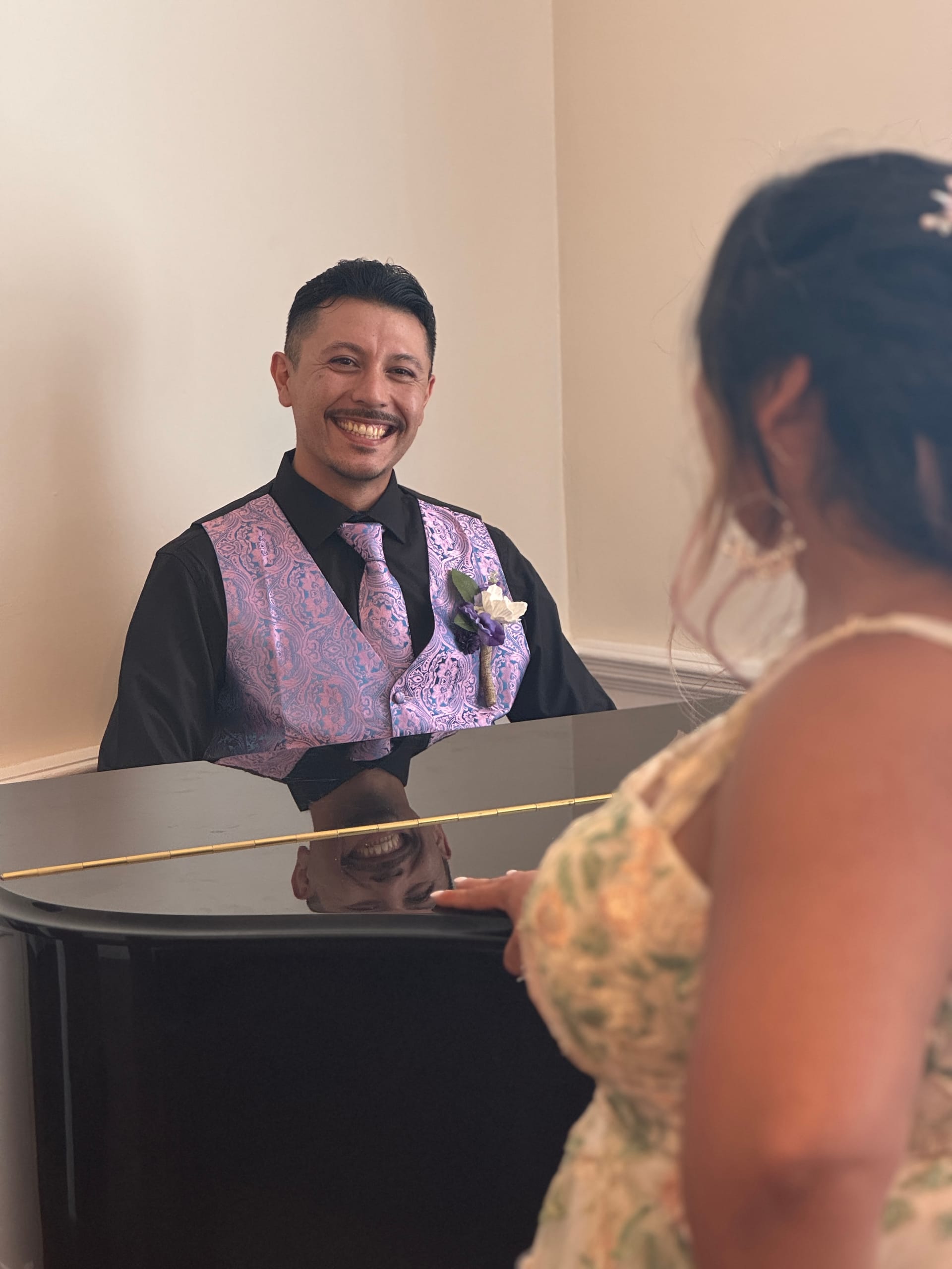 Smiling groom in a purple paisley vest and tie seated at a black grand piano sharing a joyful moment with his bride at Scarborough House.