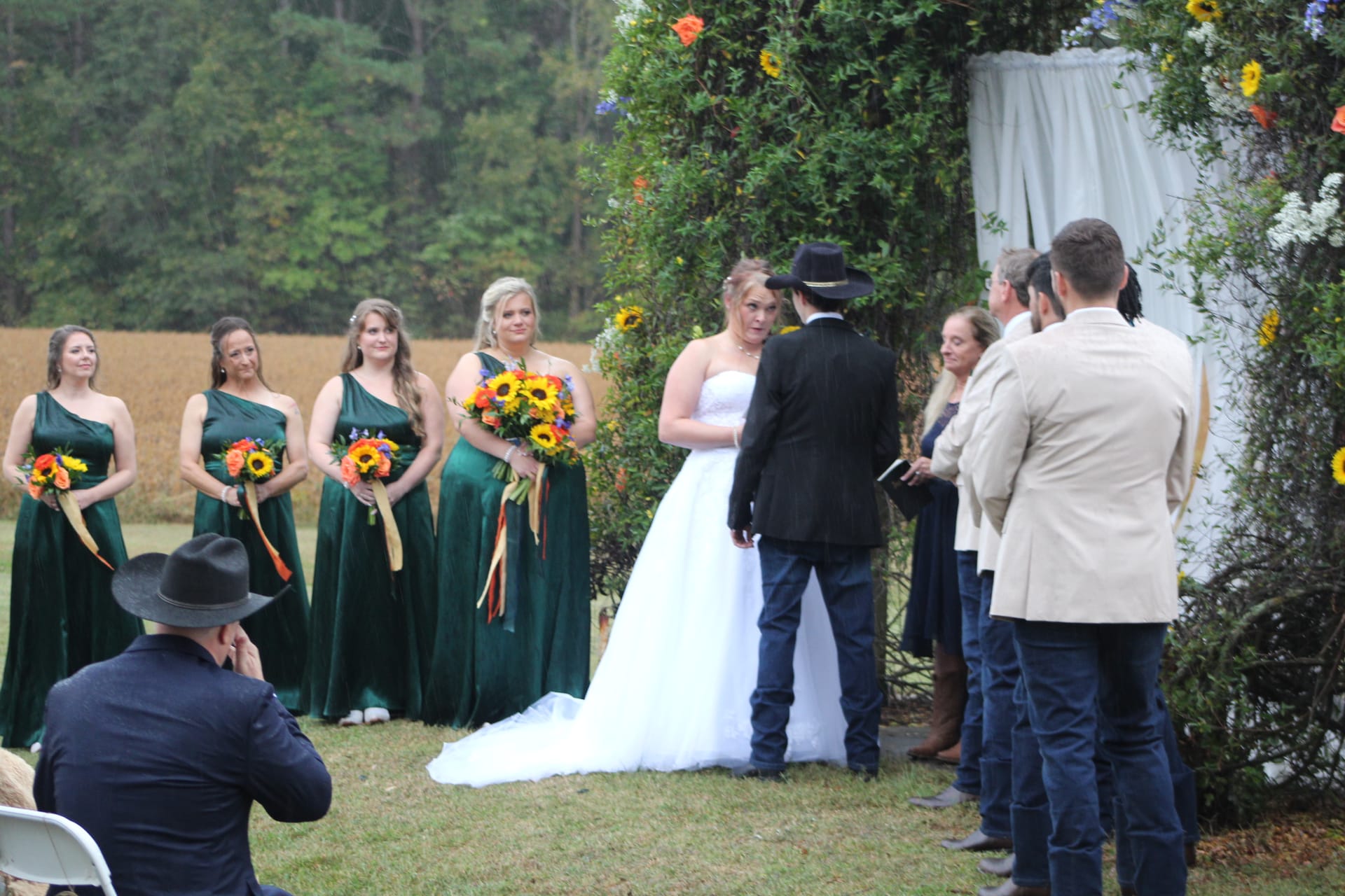 Bride and groom exchange vows at a floral arch on the ceremony lawn while four bridesmaids in emerald green gowns hold vibrant sunflower bouquets at Scarborough House.