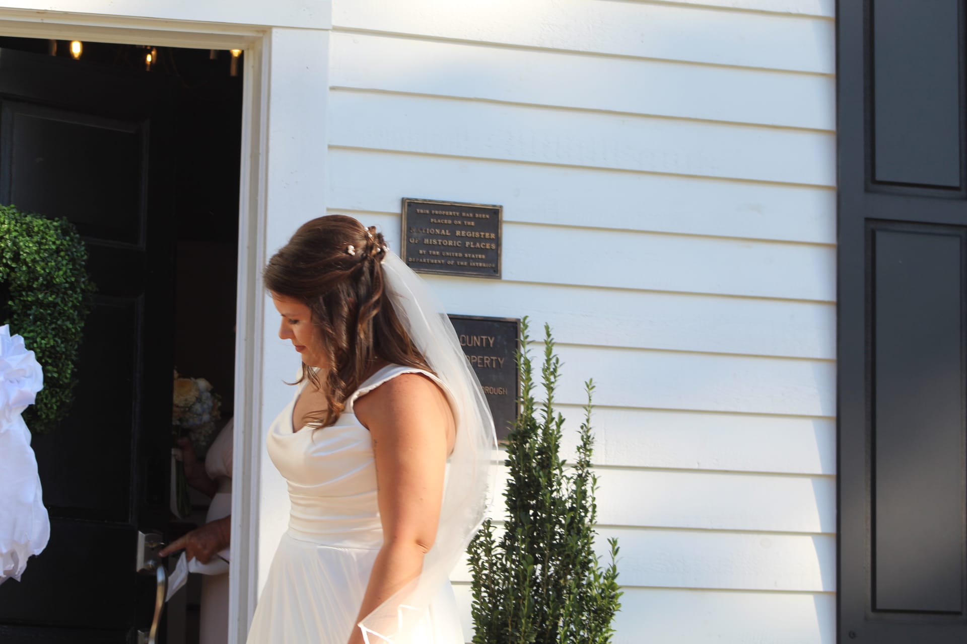 Bride in a flowing white gown and veil pausing outside the historic 1821 white clapboard manor at Scarborough House, with the National Register of Historic Places plaque visible on the wall behind her.