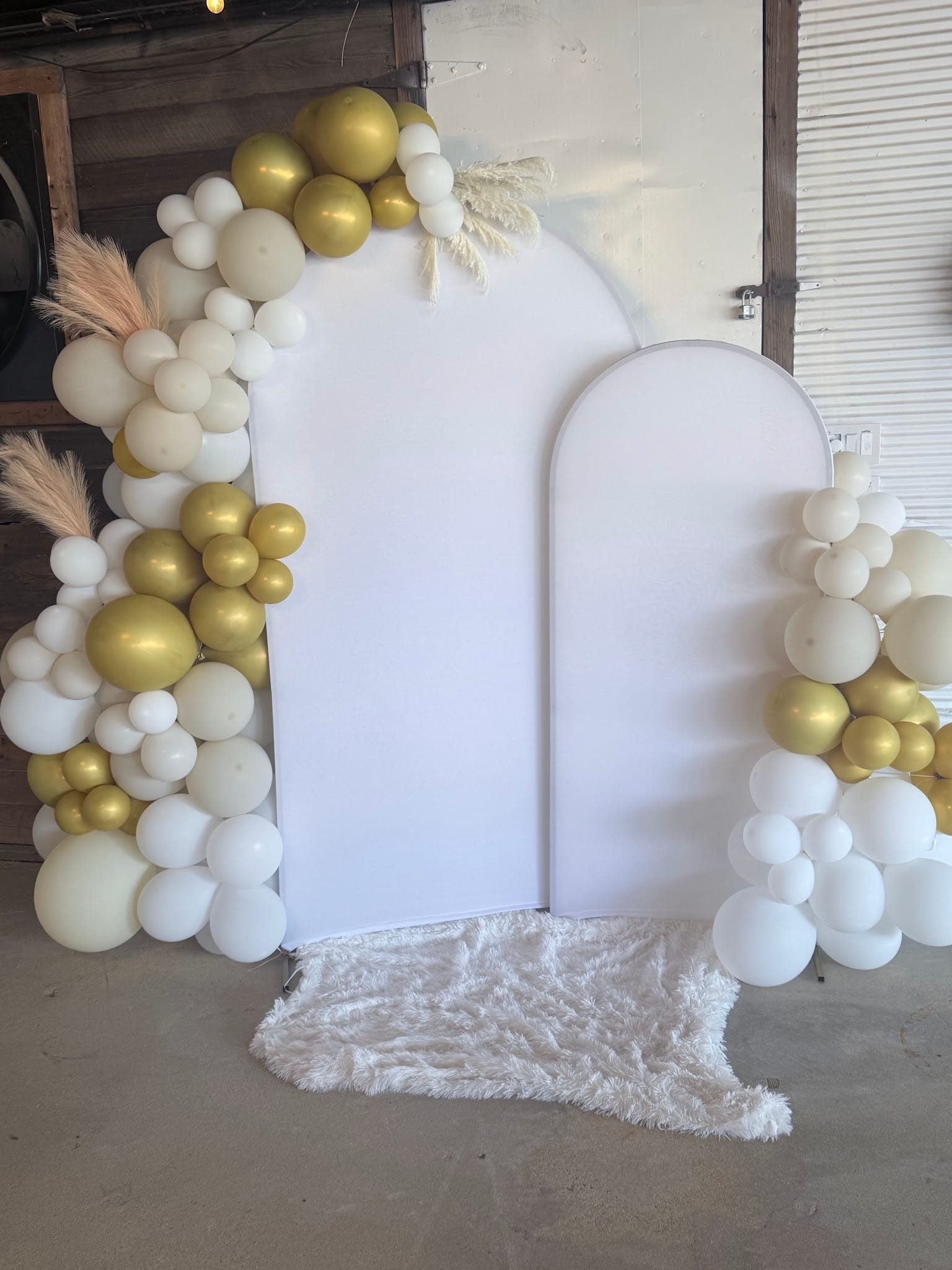 Elegant white arch backdrop flanked by organic balloon garlands in gold, ivory, and white with pampas grass accents on a fluffy white rug inside a venue event space.