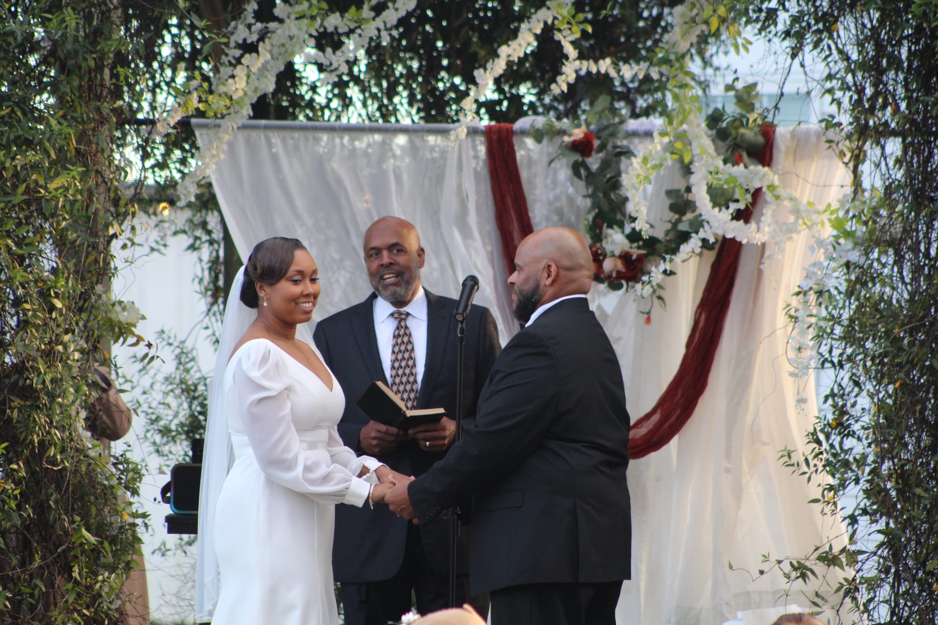 Bride in a long-sleeve white gown smiles joyfully while exchanging vows with her groom beneath a floral-adorned arch with white and burgundy drapery at Scarborough House.