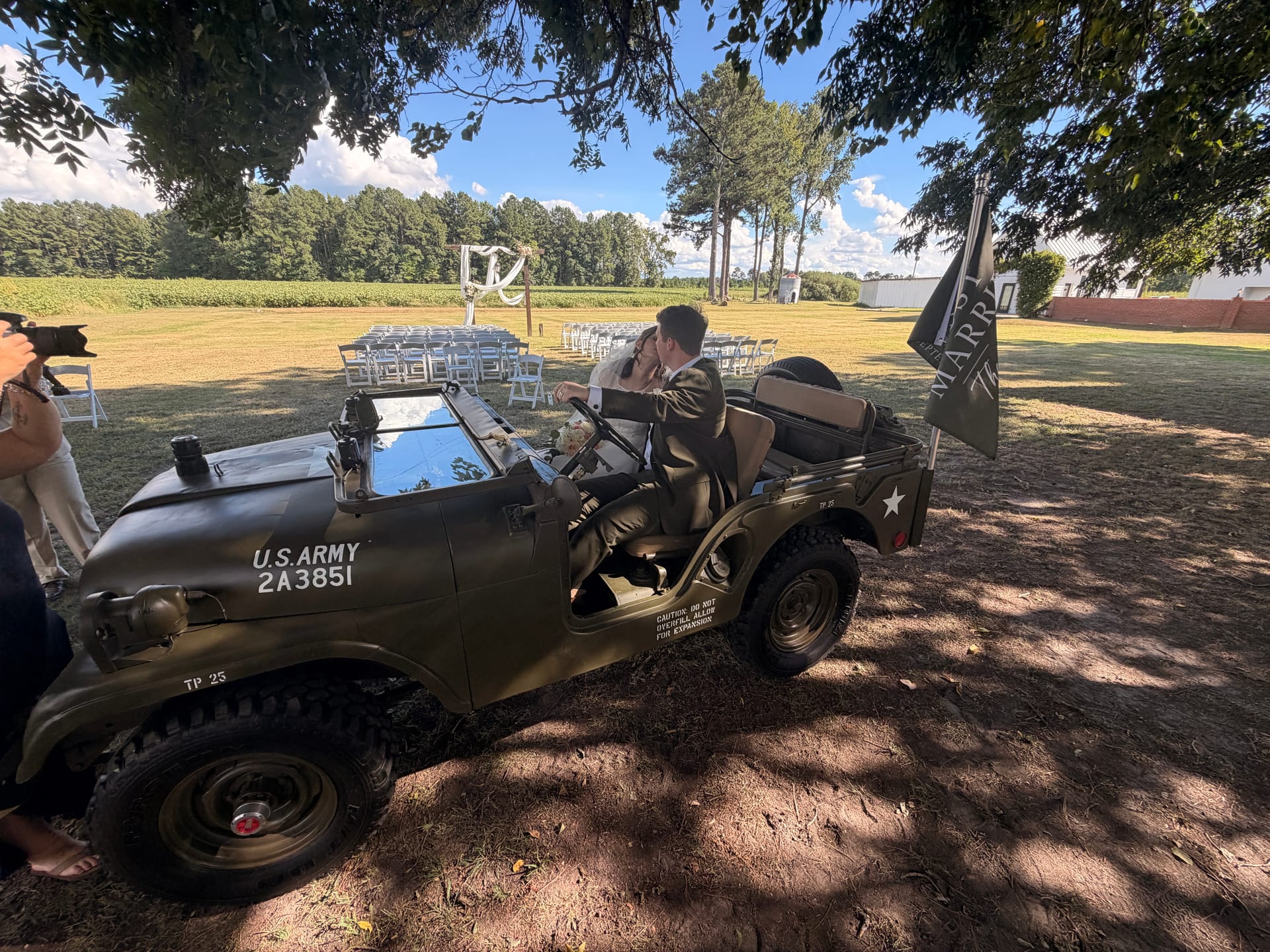 Newlyweds sharing a kiss in a vintage U.S. Army jeep adorned with a Just Married flag on the ceremony lawn at Scarborough House, with white folding chairs and a rustic outdoor altar visible in the background.