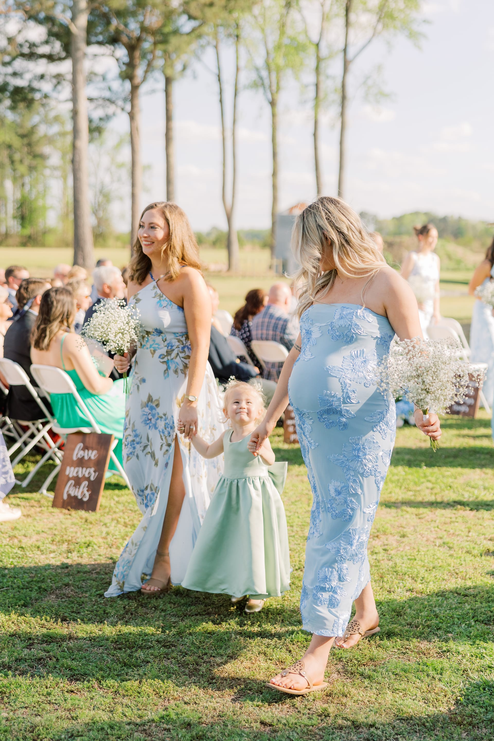 Two bridesmaids in floral and blue dresses walk down the outdoor ceremony aisle holding baby's breath bouquets while escorting a smiling flower girl in a green dress.