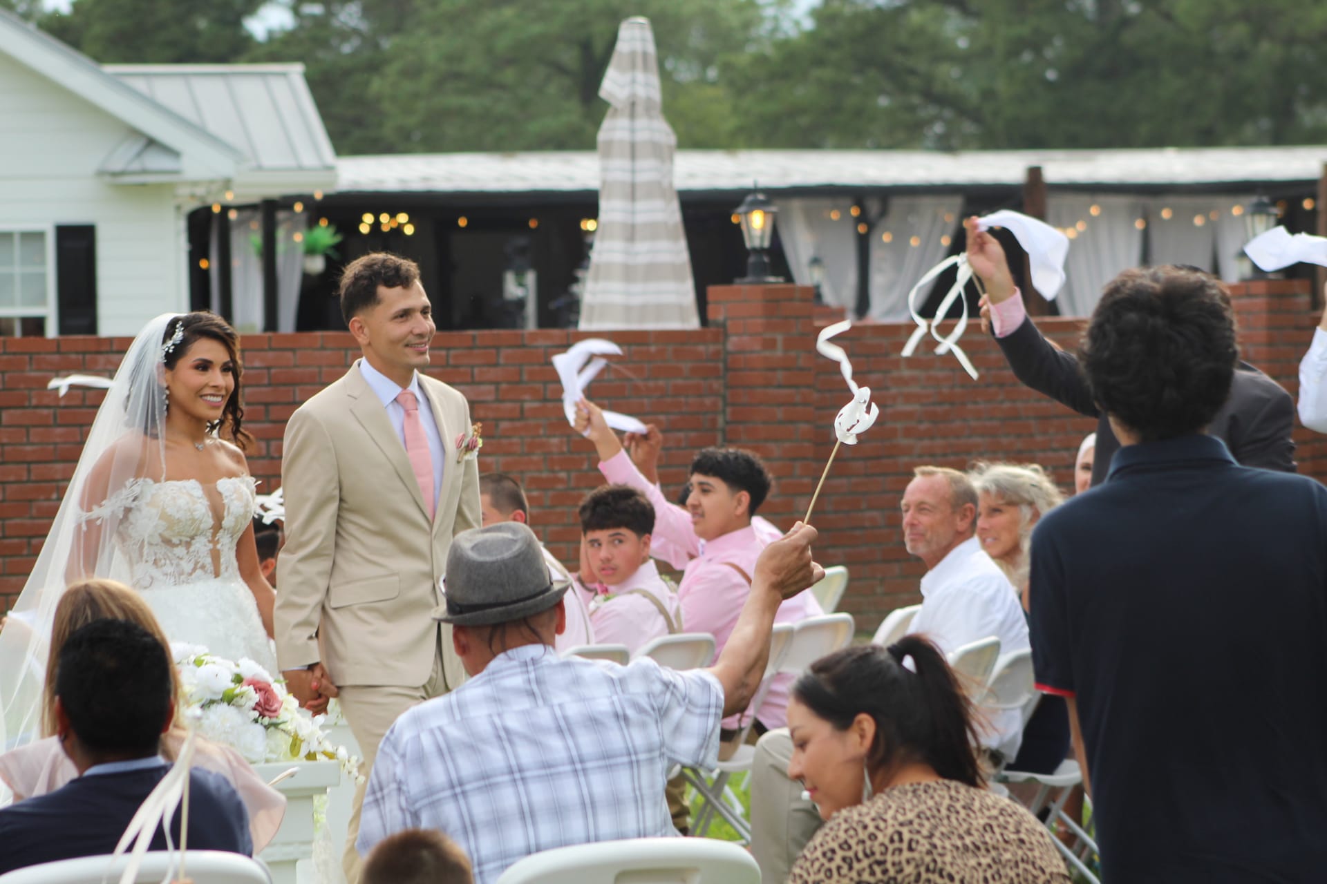 Bride in lace gown and groom in tan suit smile as guests wave white ribbon wands during their outdoor wedding recessional at a brick-walled garden venue.