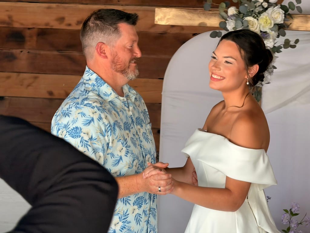 A smiling bride in an off-shoulder white gown holds hands with the groom beneath a floral arch during their wedding ceremony at Scarborough House.