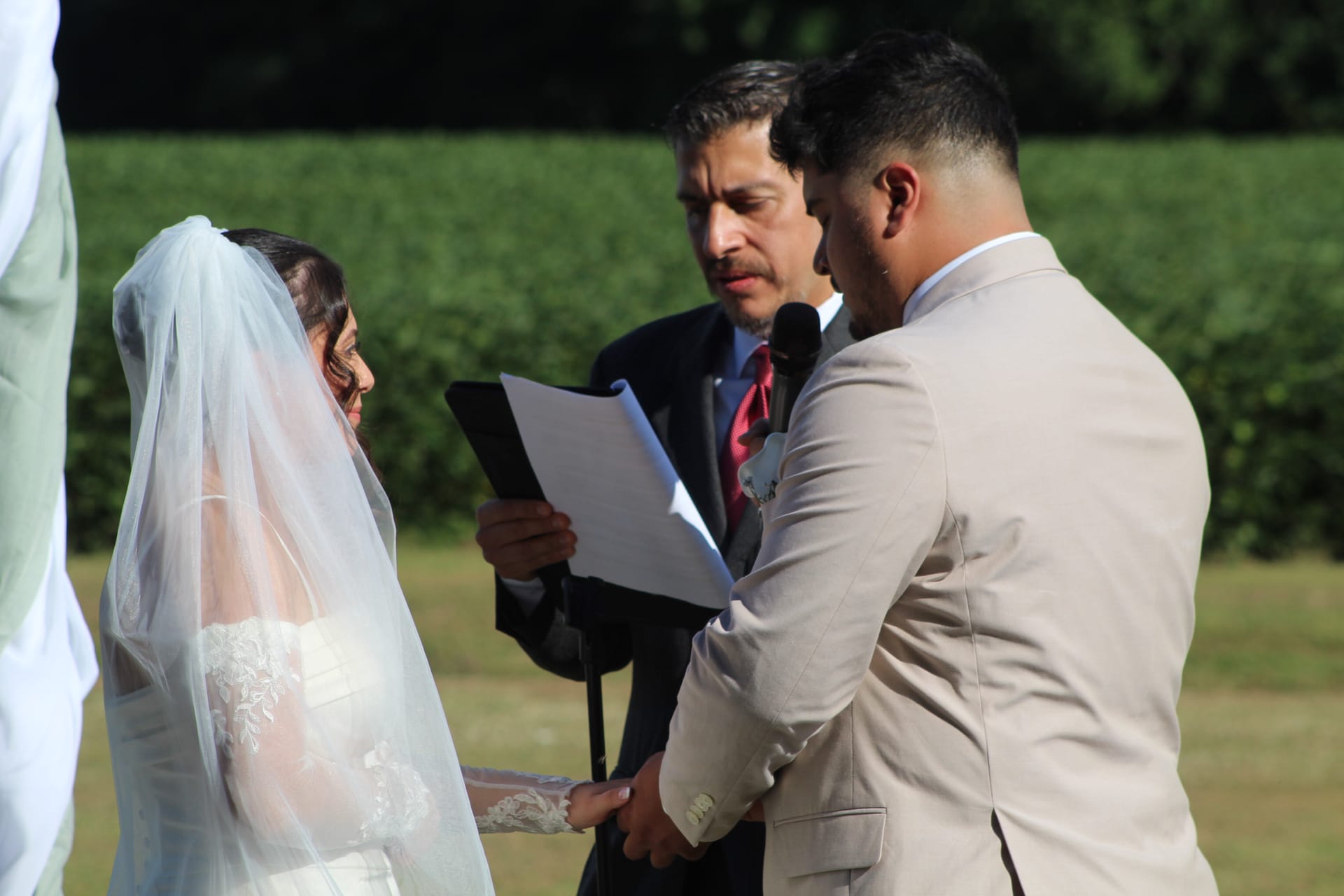 Bride and groom hold hands during outdoor vow exchange as officiant reads from ceremony papers on the sunlit ceremony lawn at Scarborough House.