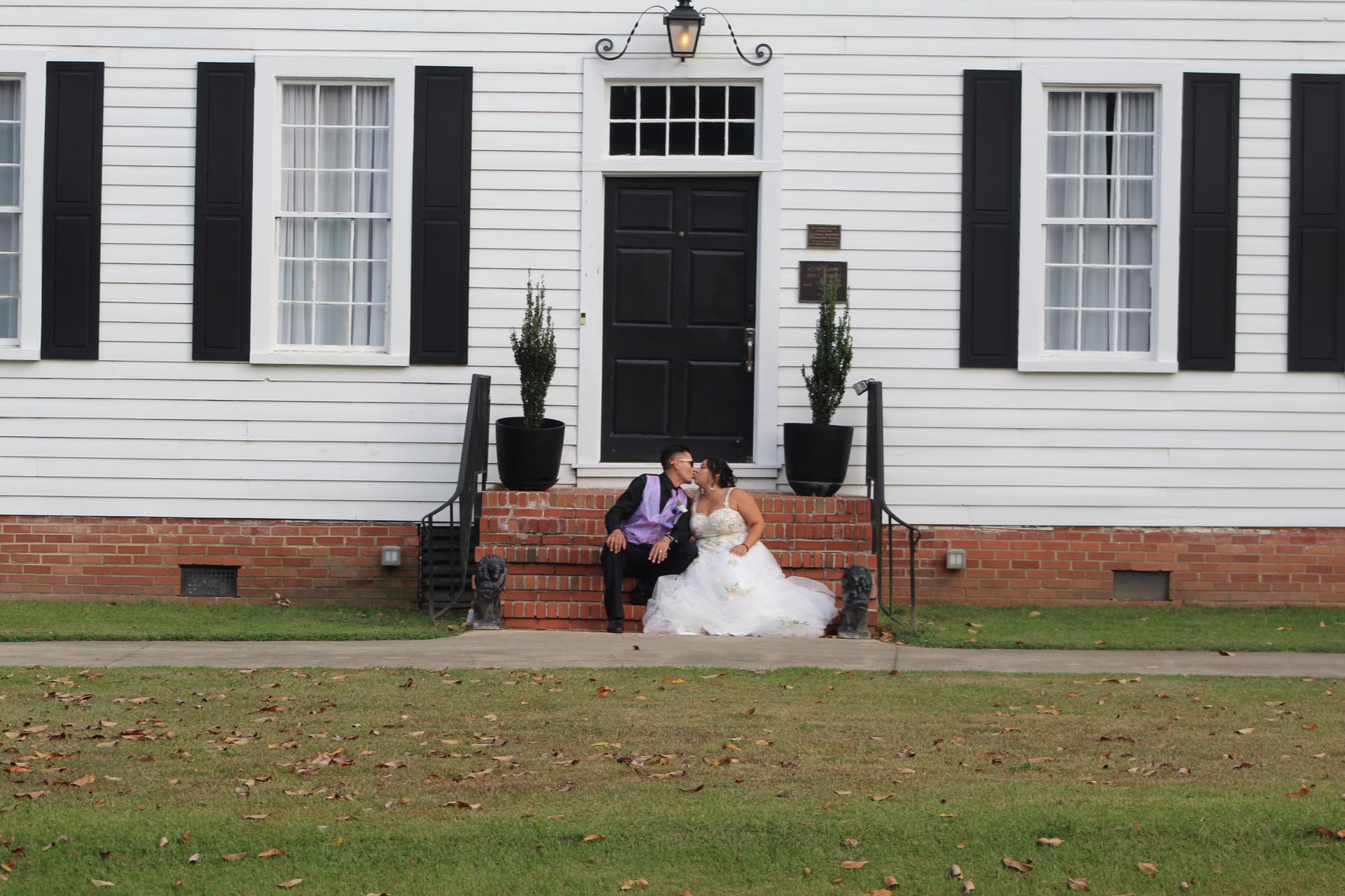 Bride and groom share a kiss while seated on the brick front steps of the 1821 Scarborough House manor, framed by black shutters and potted topiaries.