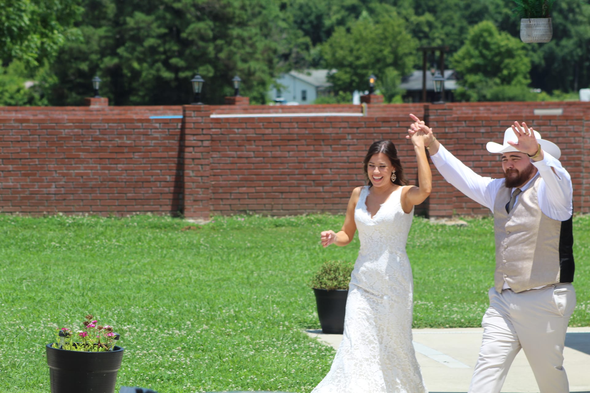 Bride in white lace gown and groom in tan vest and cowboy hat celebrate their grand entrance on the sunlit ceremony lawn at Scarborough House.