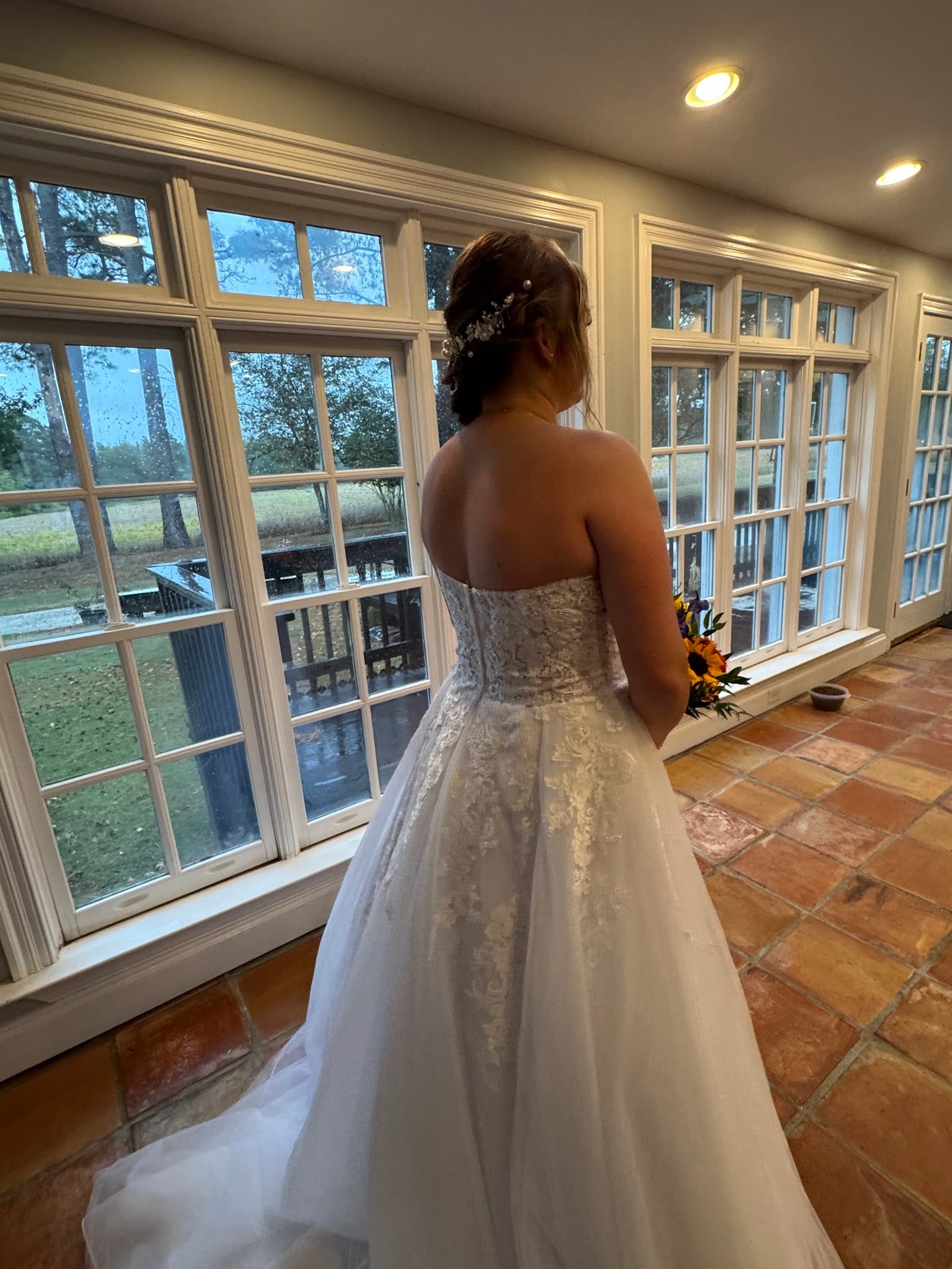 Bride in a strapless lace wedding gown holding a sunflower bouquet gazes out large white-framed windows at the historic Scarborough House estate on a rainy wedding day.