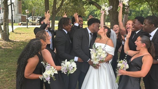 Bride and groom share a kiss surrounded by their cheering wedding party dressed in black beneath lush oak trees at Scarborough House.