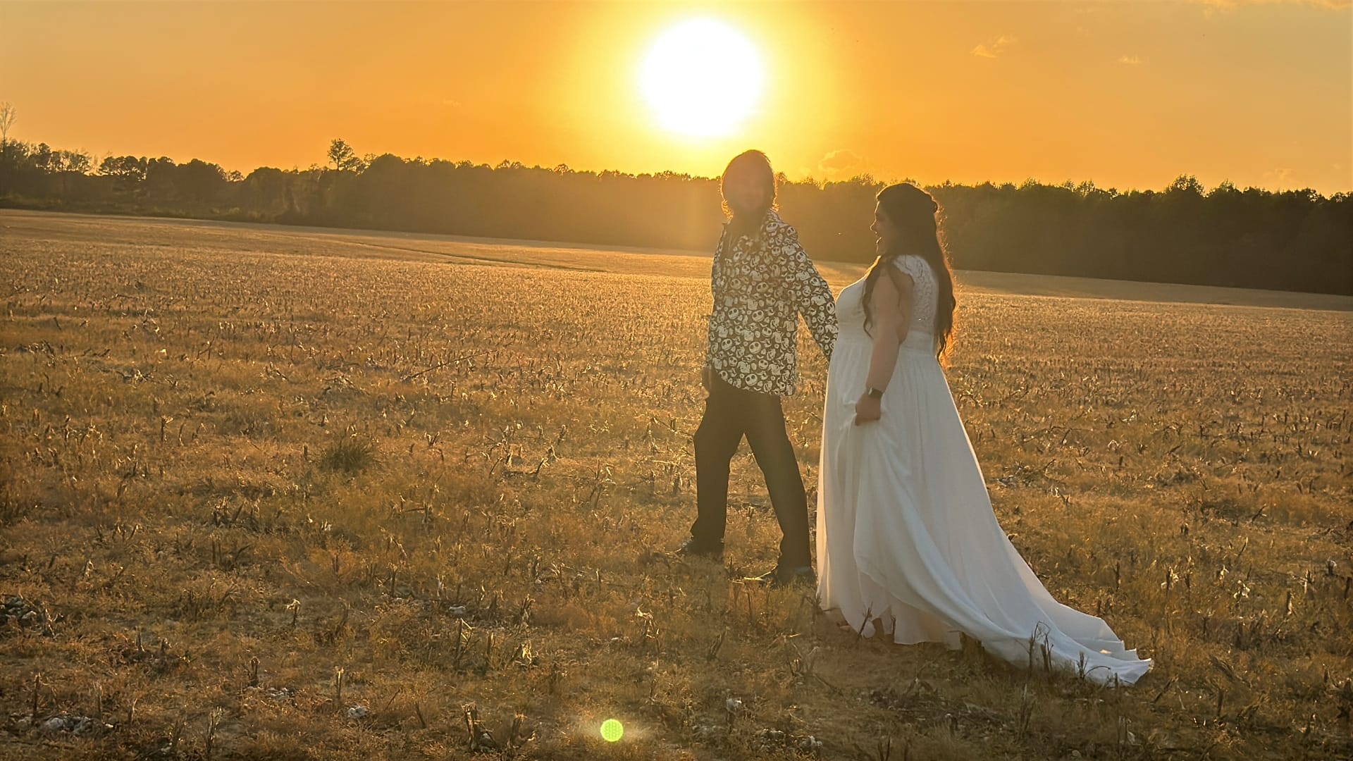 Bride in flowing white gown and groom in floral shirt walk hand in hand through golden farmland fields at sunset at Scarborough House estate wedding venue.