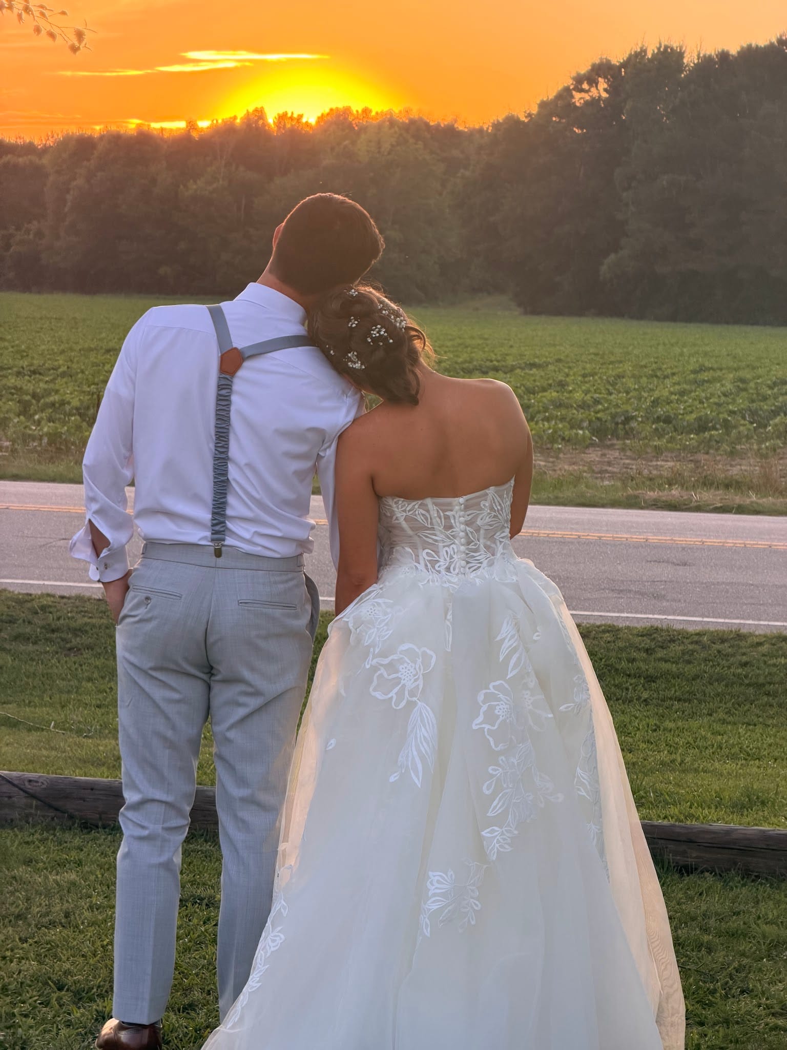 Bride in a floral lace ball gown leans her head on the groom's shoulder as they watch a vivid orange sunset over open farmland fields at Scarborough House.