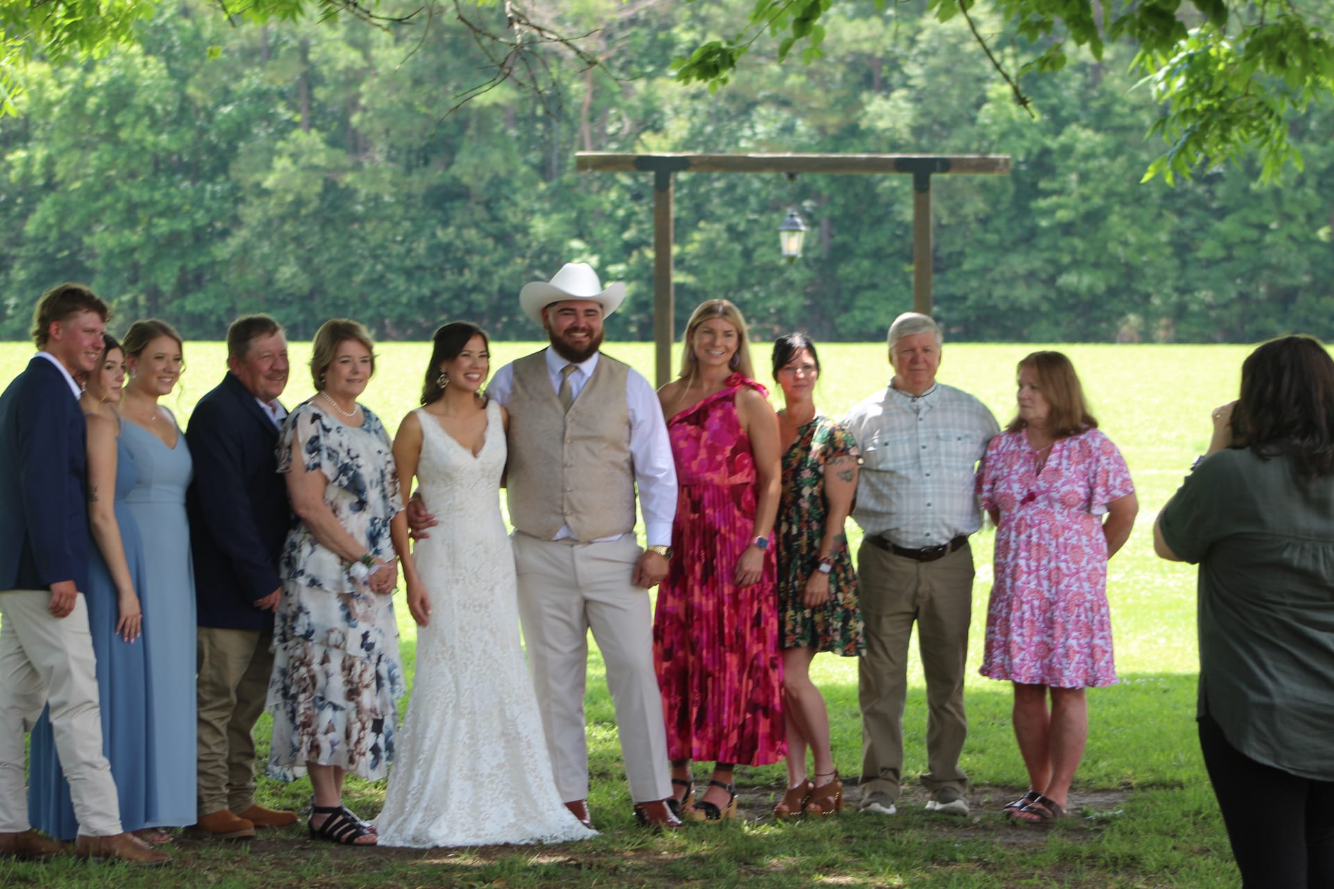 Bride in white lace gown and groom in tan suit and white cowboy hat pose with family members on the ceremony lawn at Scarborough House, surrounded by lush green trees and a rustic wooden arbor.