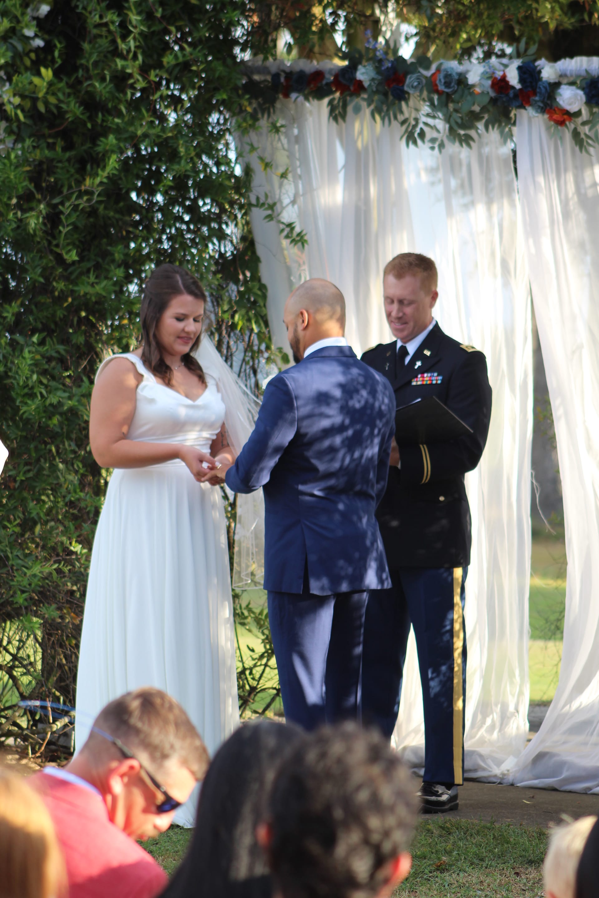 Bride in a white gown exchanges rings with her groom beneath a draped floral arch adorned with red, white, and blue flowers as a military officiant looks on during an outdoor wedding ceremony.