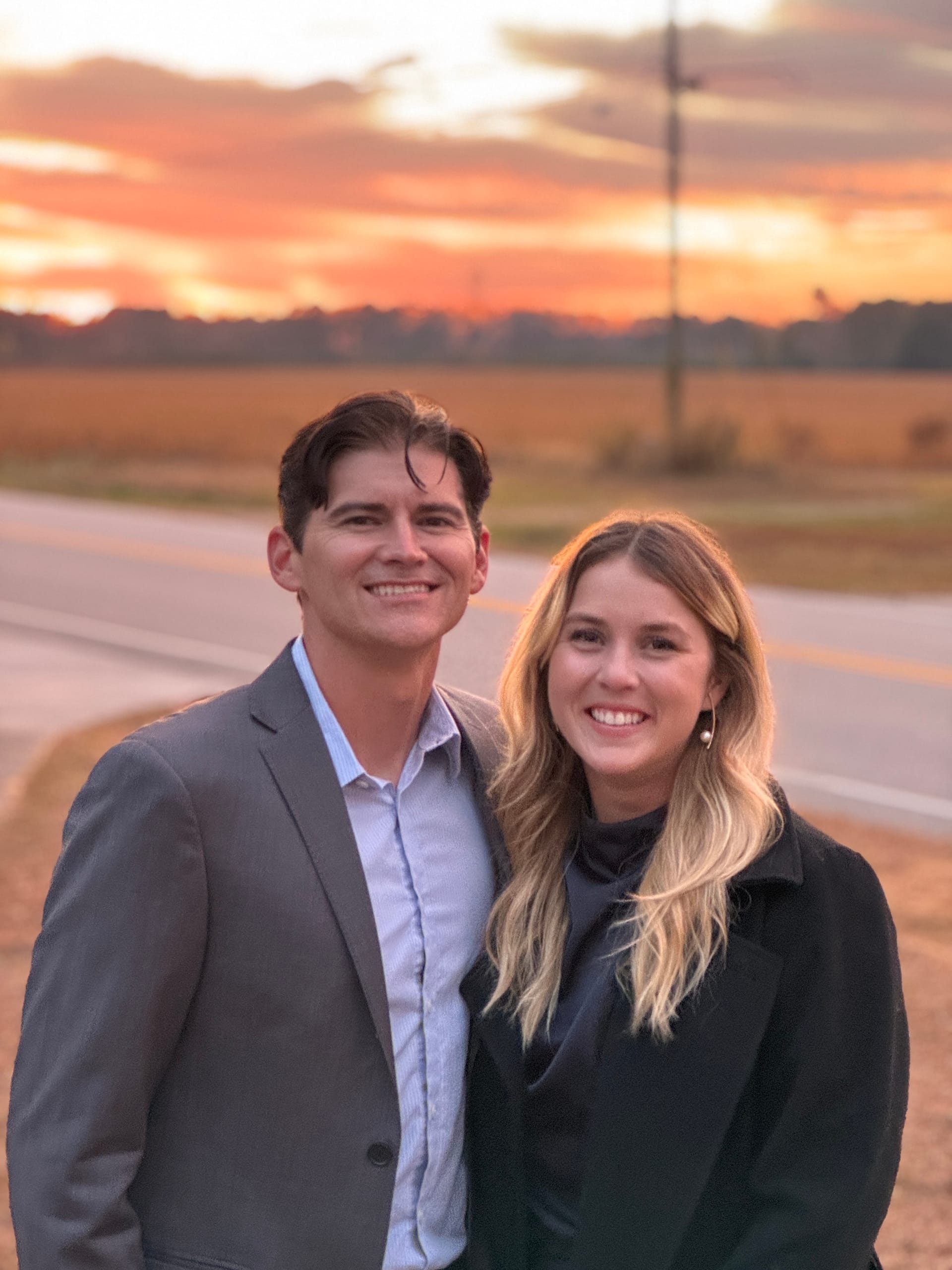 A smiling couple poses together in front of a vibrant orange and pink sunset sky along the open farmland fields at Scarborough House.