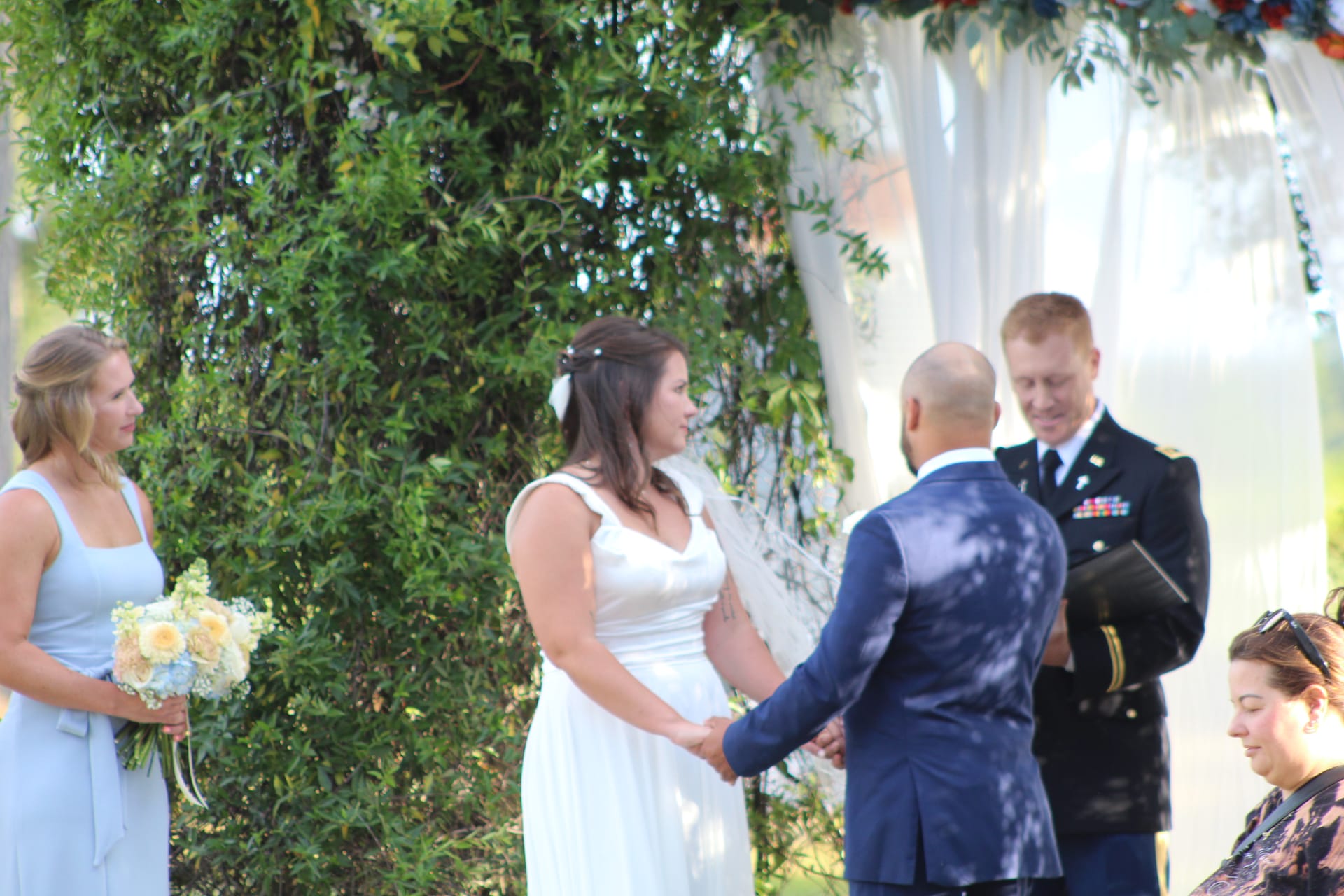 Bride in white gown holds hands with groom during outdoor wedding ceremony officiated by a military officer beneath a floral arch at Scarborough House.