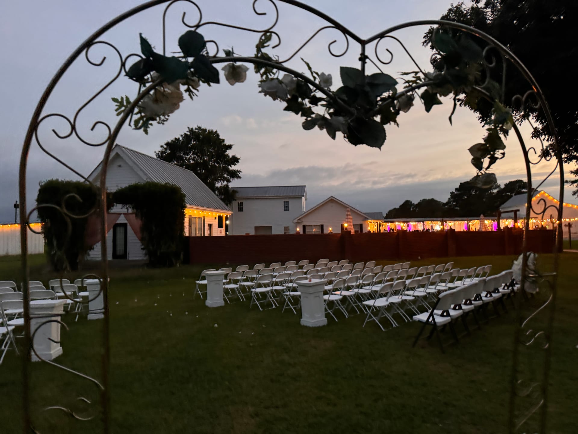 Outdoor wedding ceremony setup at dusk viewed through a heart-shaped wrought-iron arch adorned with white flowers, with rows of white folding chairs on the lawn and a warmly lit reception pavilion glowing in the background at Scarborough House.