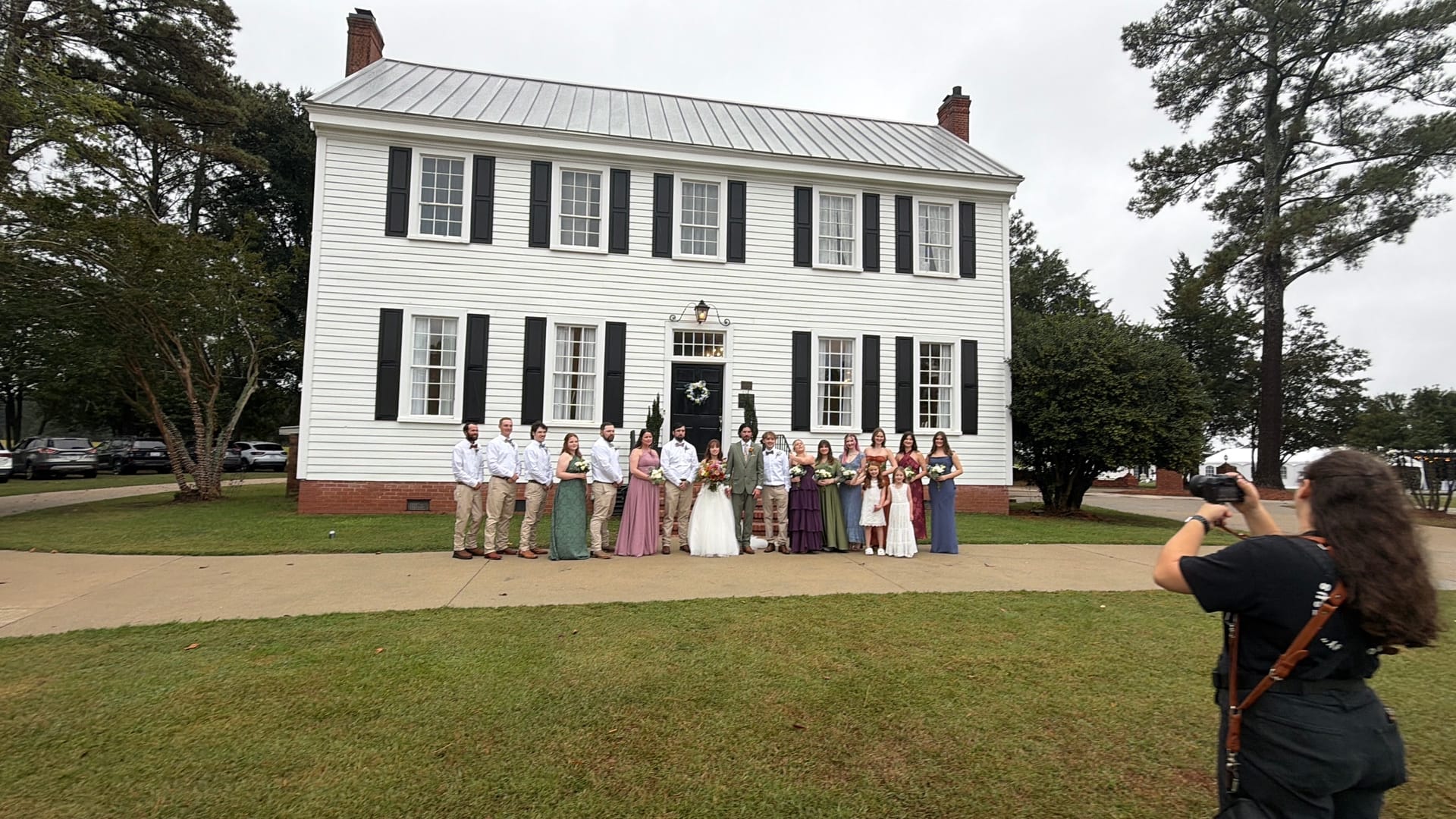 Full wedding party posing in front of the white 1821 Scarborough House manor with black shutters while a photographer captures the group portrait on the ceremony lawn.