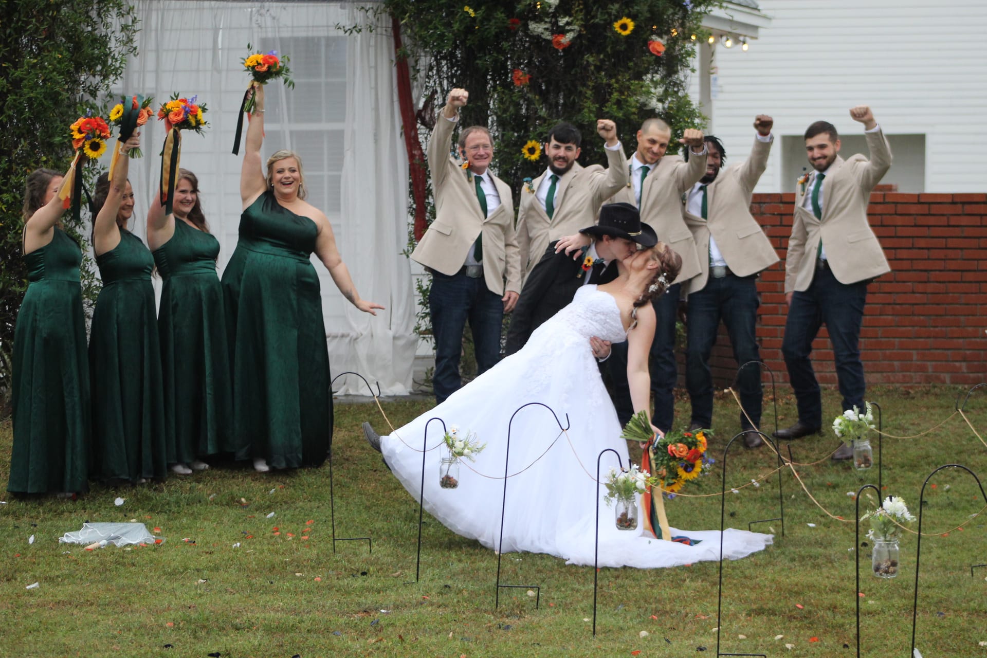 Bride and groom share a dramatic dip kiss on the ceremony lawn at Scarborough House while bridesmaids in emerald gowns and groomsmen in tan suits cheer and raise their fists in celebration.