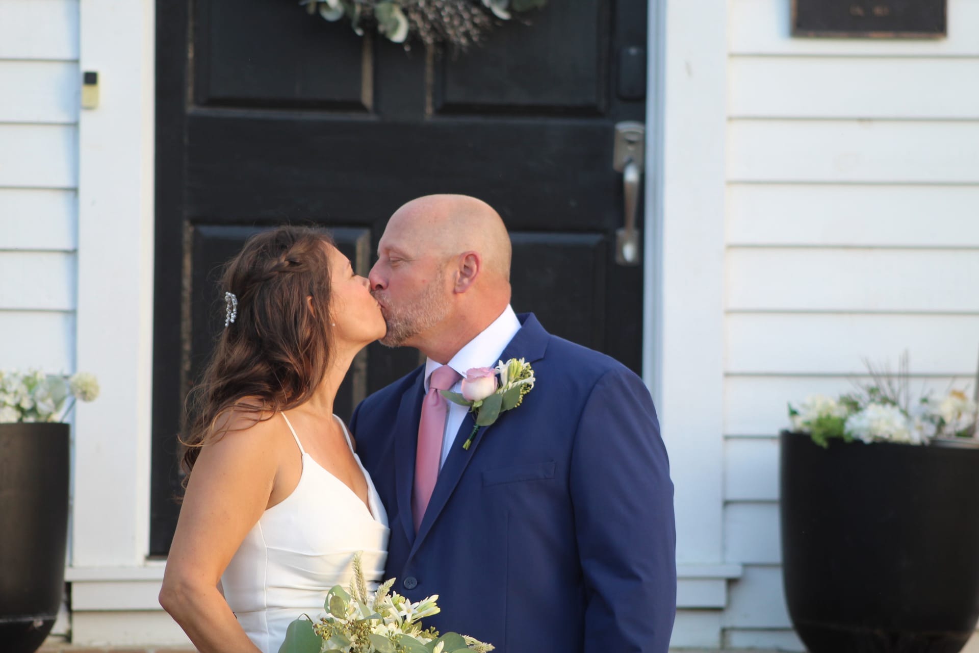 Bride in a white spaghetti-strap gown and groom in a navy suit with pink tie share a kiss in front of the black door of the 1821 Scarborough House manor.