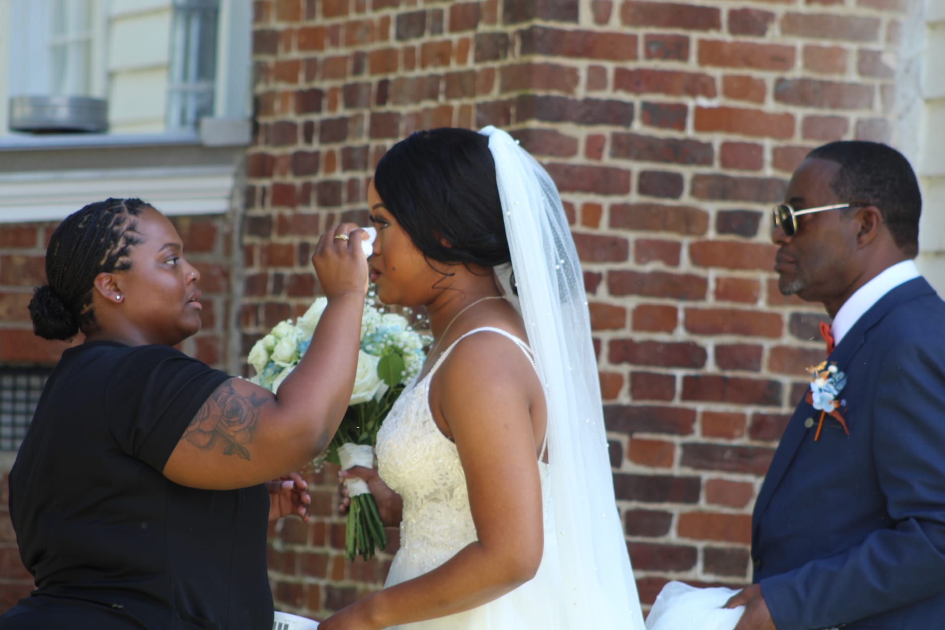A bridesmaid dabs tears from an emotional bride's eye as the groom looks on tenderly outside a historic brick manor wedding venue.