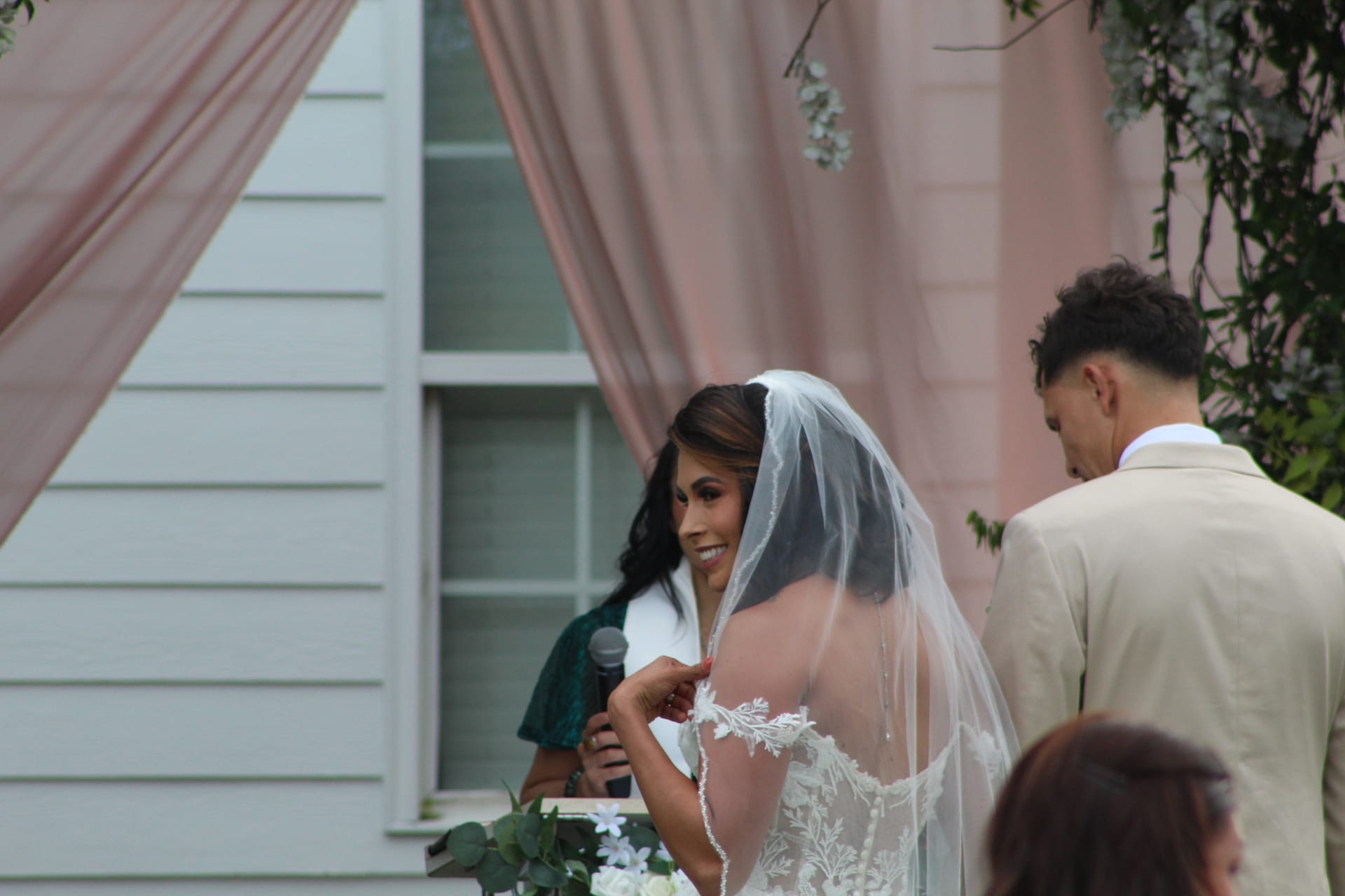 Smiling bride in lace gown and veil exchanges vows with groom in tan suit during an outdoor wedding ceremony beneath blush pink drapery at Scarborough House.