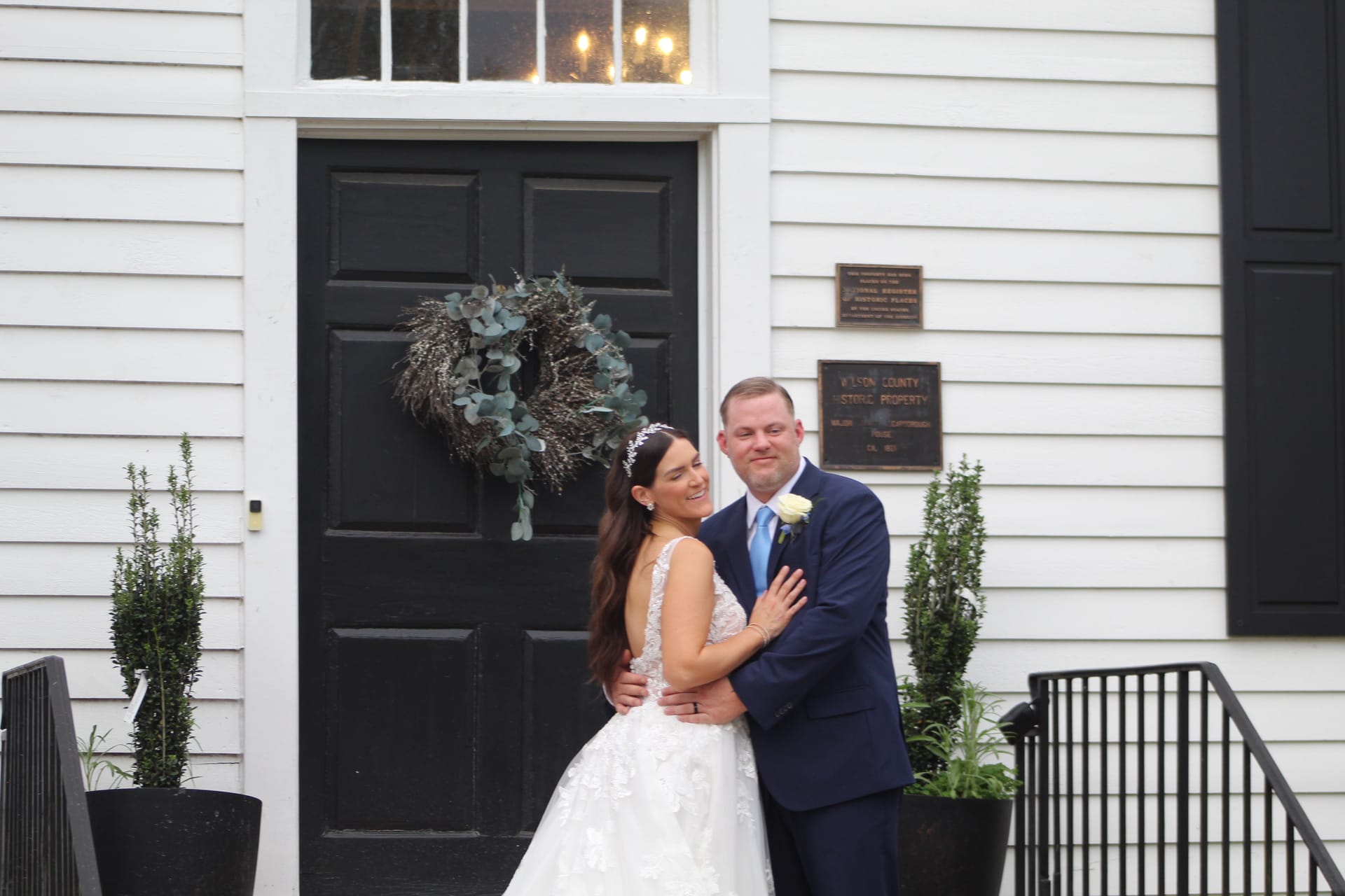 Bride in lace gown and groom in navy suit embracing and smiling in front of the black-doored entrance of the 1821 Scarborough House historic manor, adorned with a eucalyptus wreath.