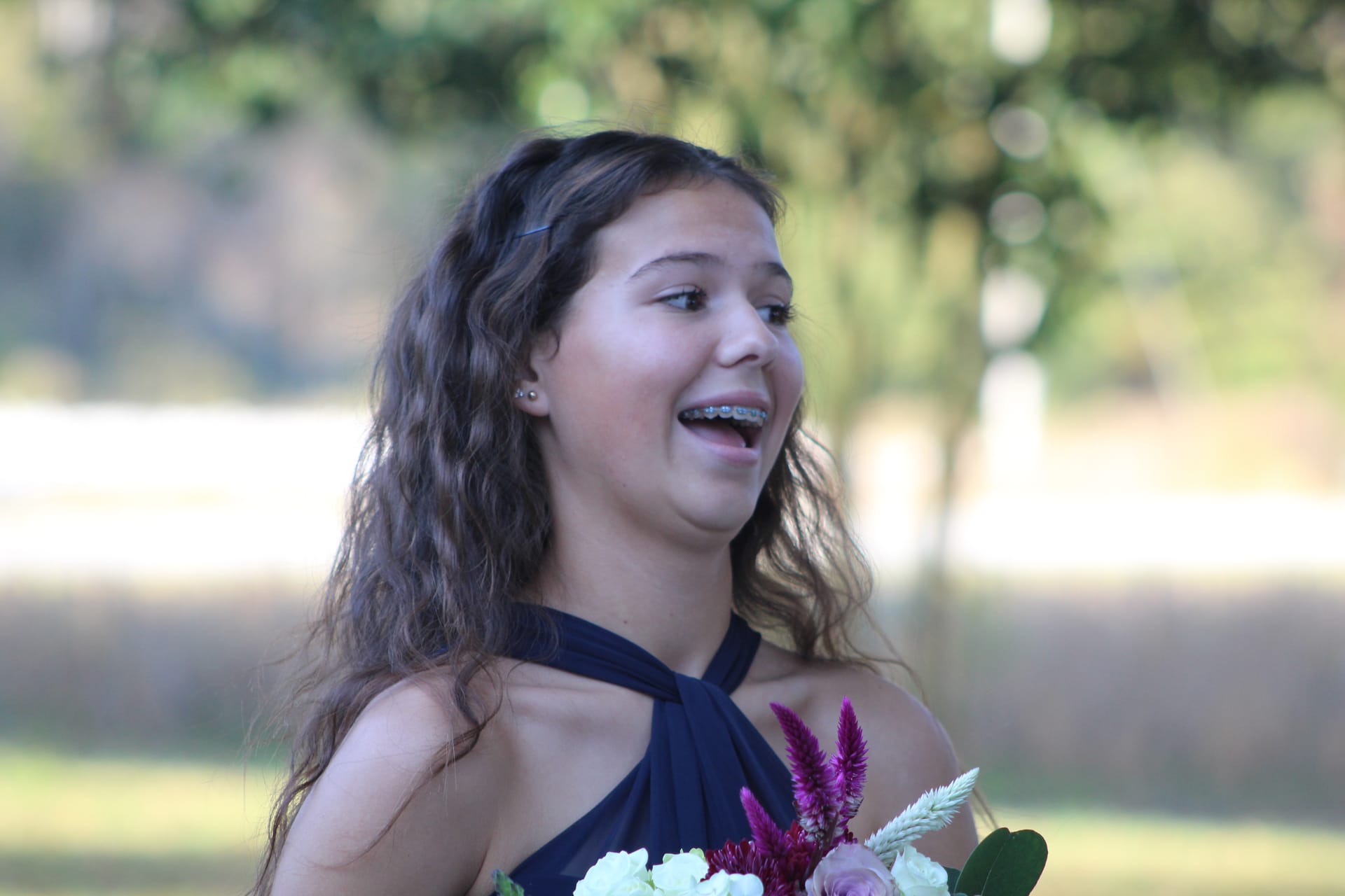 A smiling young bridesmaid in a navy halter dress holds a bouquet of white, pink, and magenta flowers on the ceremony lawn surrounded by blurred oak trees and open fields.