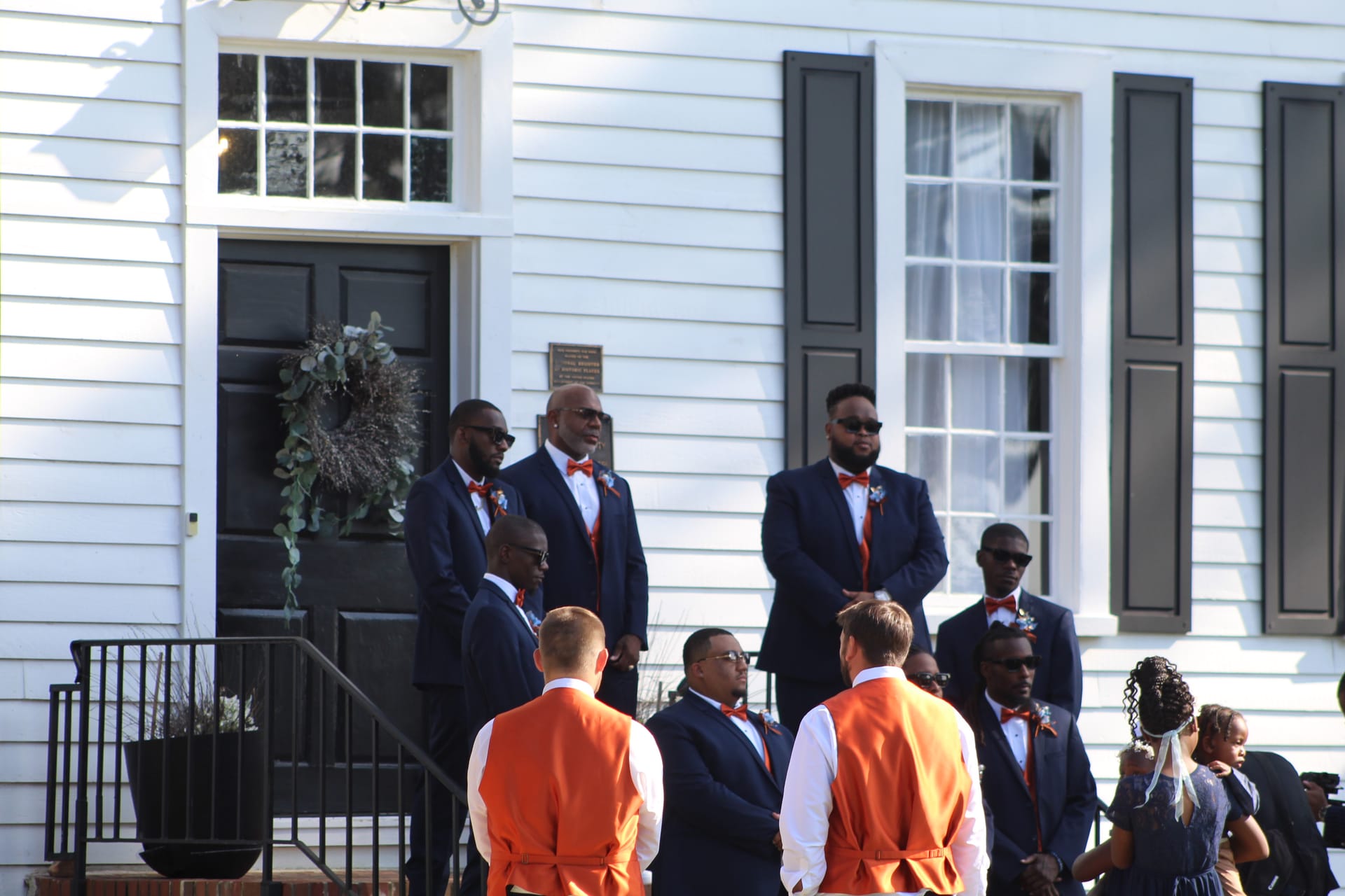 Groomsmen in navy suits with orange bow ties and vests gathered on the front steps of the 1821 Scarborough House manor during a wedding ceremony.