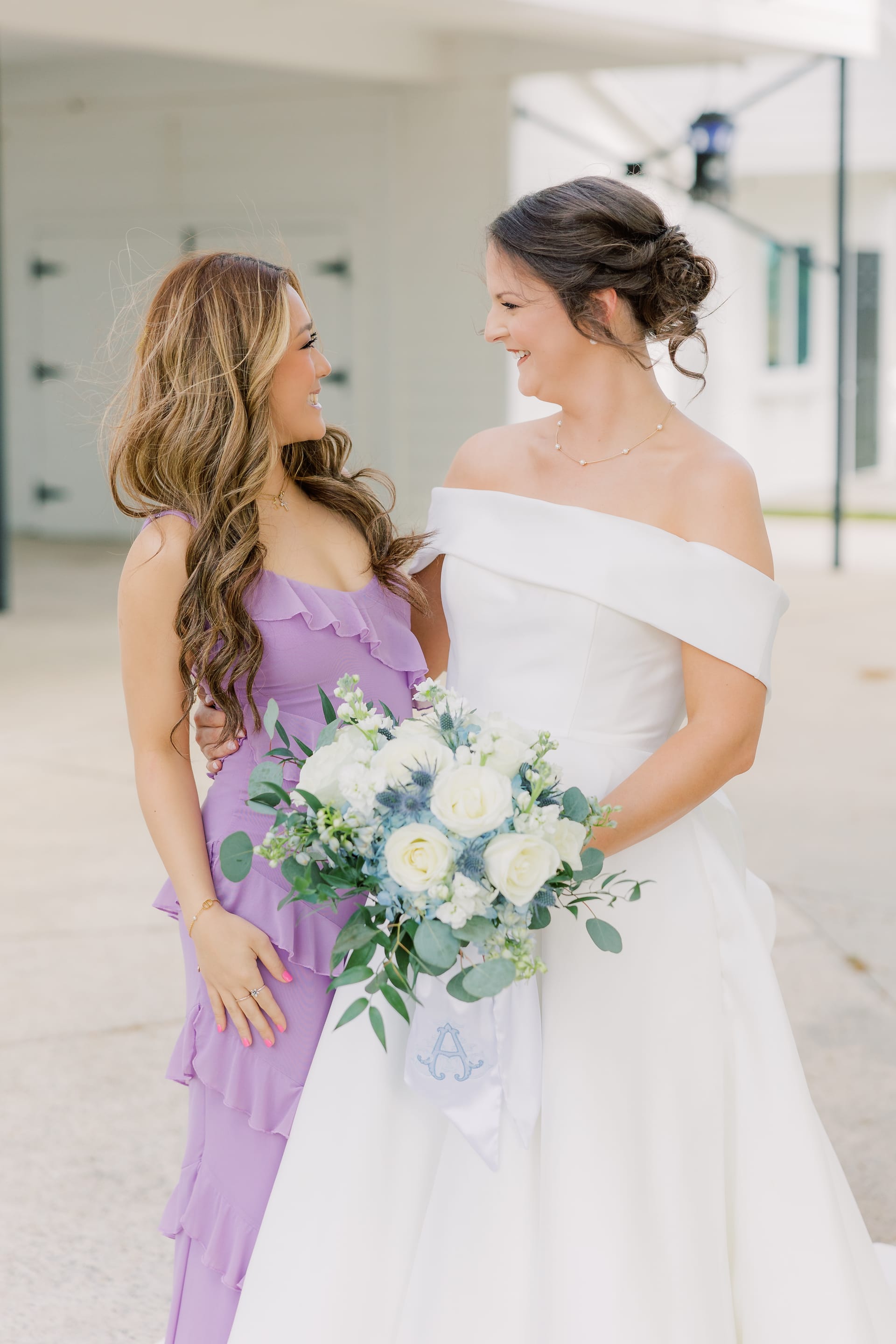 Smiling bride in an off-the-shoulder white gown holding a white rose and eucalyptus bouquet stands beside a bridesmaid in a lavender ruffled dress at Scarborough House.