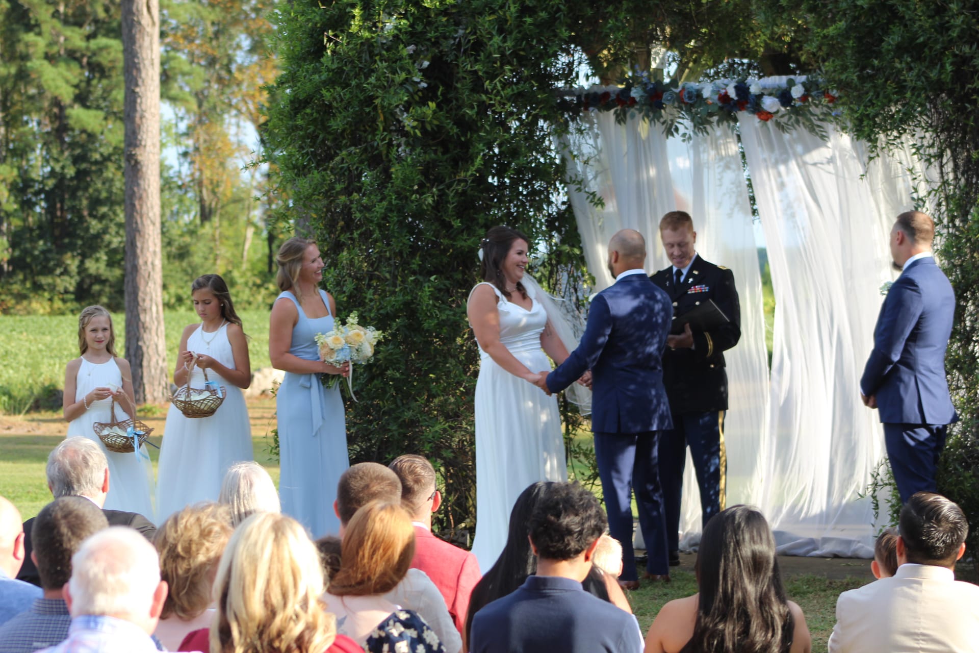 Smiling bride in white gown holds hands with groom in military dress uniform during an outdoor wedding ceremony beneath a lush floral and greenery arch draped with white fabric, flanked by bridesmaids in dusty blue and flower girls.