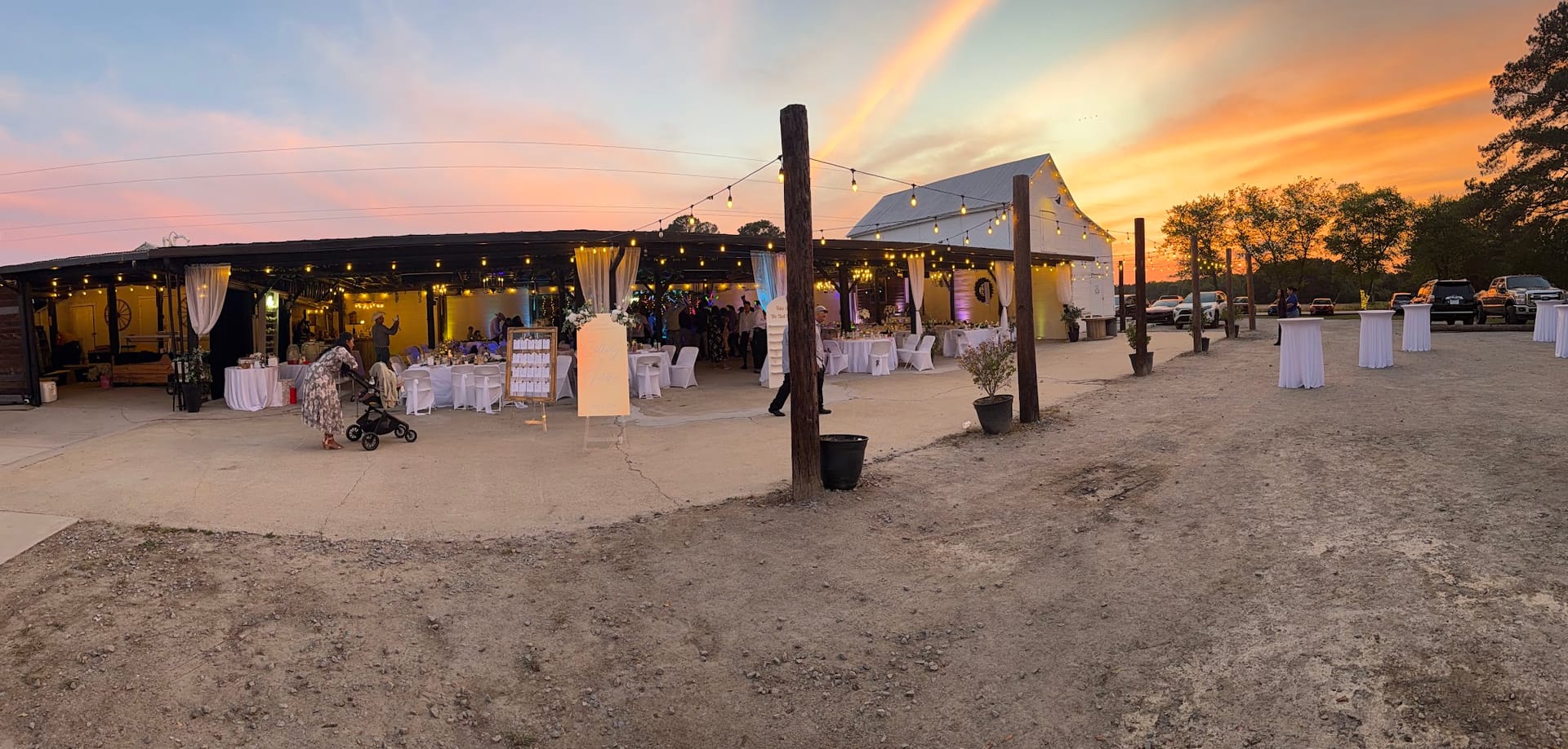 Panoramic twilight view of an outdoor wedding reception with string lights and white-draped tables glowing beneath a vivid orange and pink sunset sky.