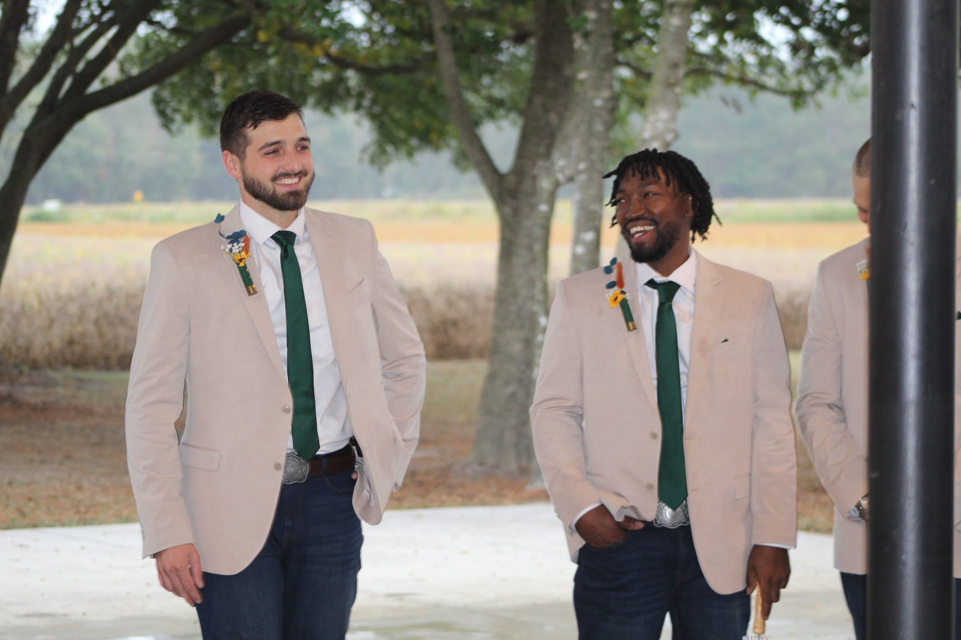 Two smiling groomsmen in tan blazers and green ties with boutonnieres share a candid laugh outdoors at Scarborough House with oak trees and open farmland fields in the background.