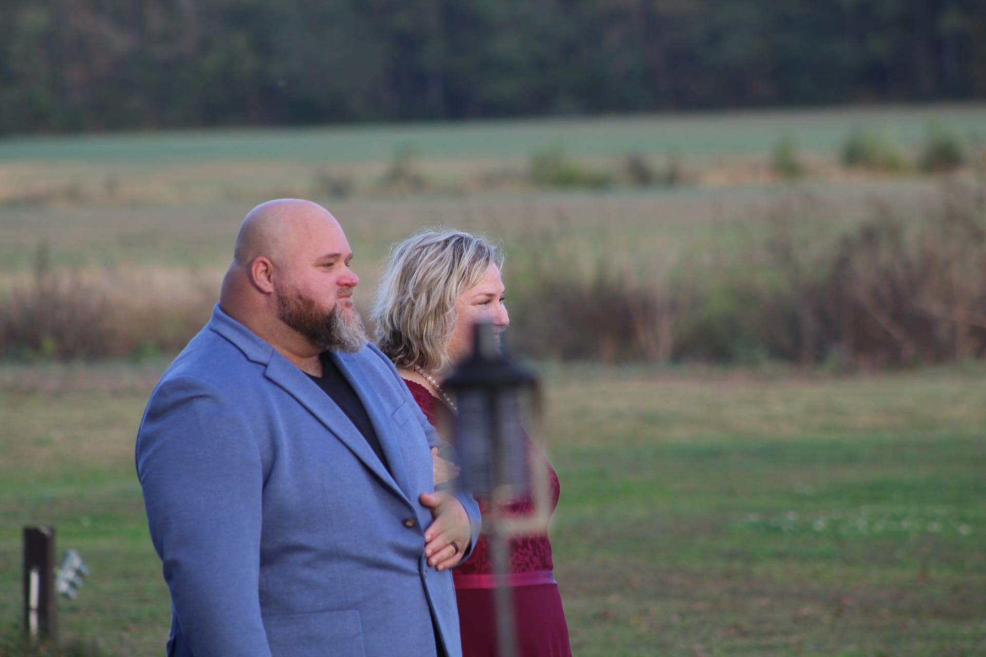 A couple dressed in a blue suit and burgundy gown stand together arm in arm watching a wedding ceremony with open farmland and tree line in the background at golden hour.