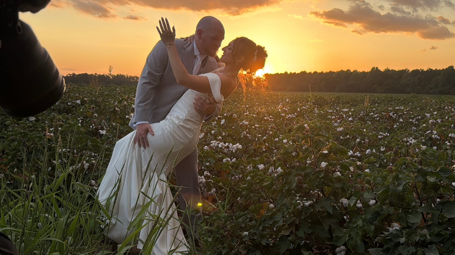 Bride and groom sharing a romantic dip kiss in a blooming cotton field at golden hour with a vivid orange sunset glowing behind them at Scarborough House.