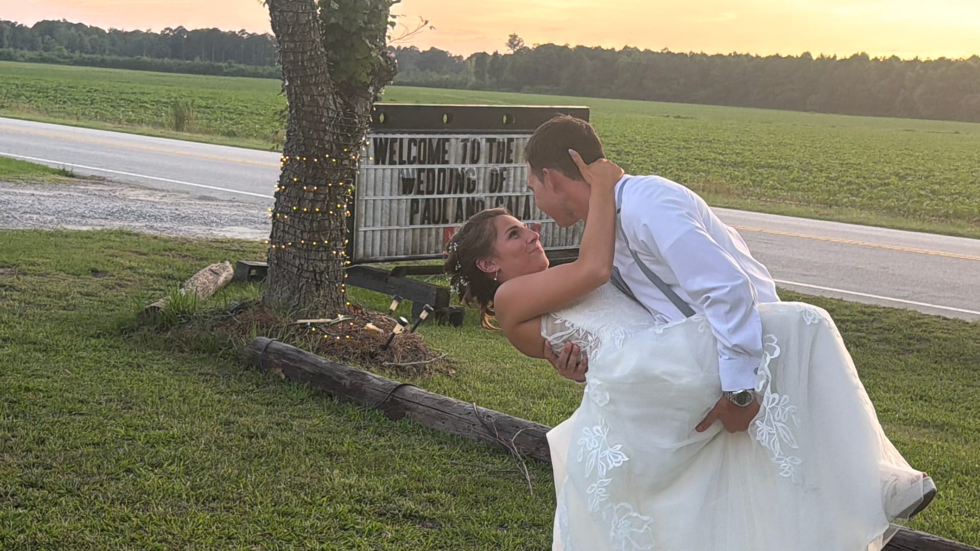 Groom dipping his bride in a romantic pose in front of the wedding welcome marquee sign at golden hour with open farmland fields and a glowing sunset sky behind them.
