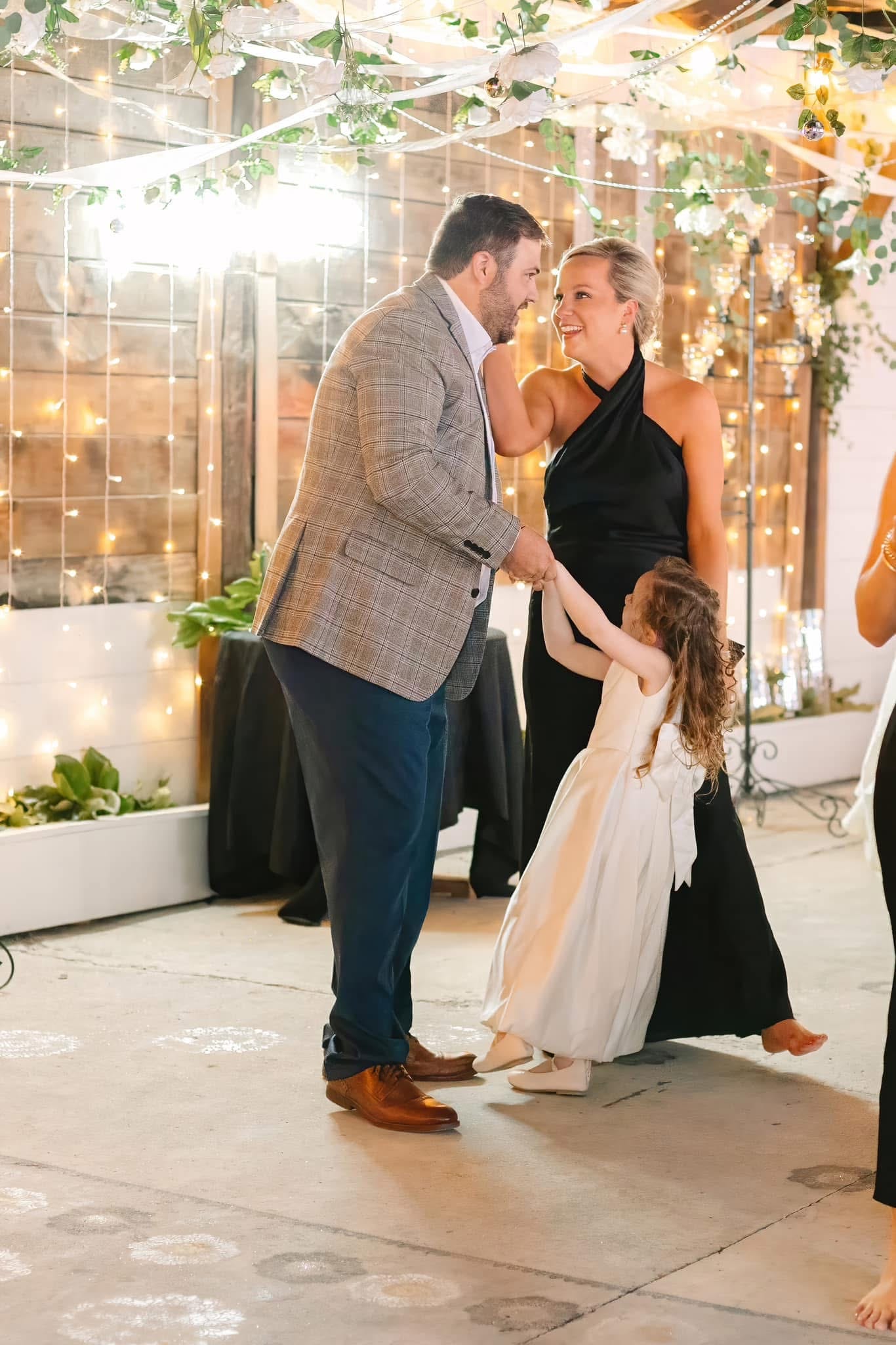 A smiling couple in formal attire dances with a young flower girl under hanging greenery and warm fairy lights at a Scarborough House wedding reception.