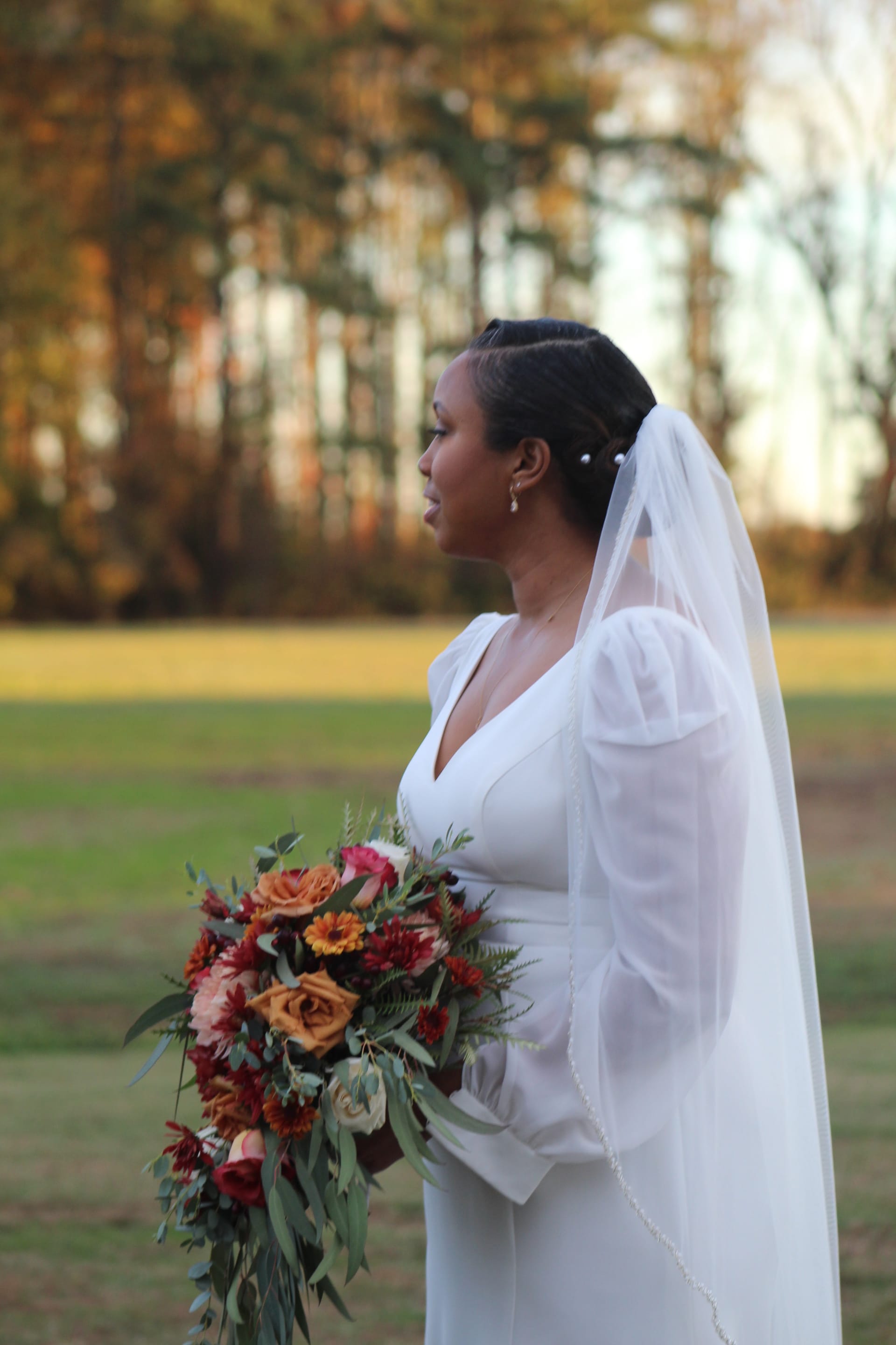 Bride in a white long-sleeve gown and veil holding a cascading autumn bouquet of burgundy, orange, and blush flowers stands in a sunlit field with fall trees in the background at Scarborough House.