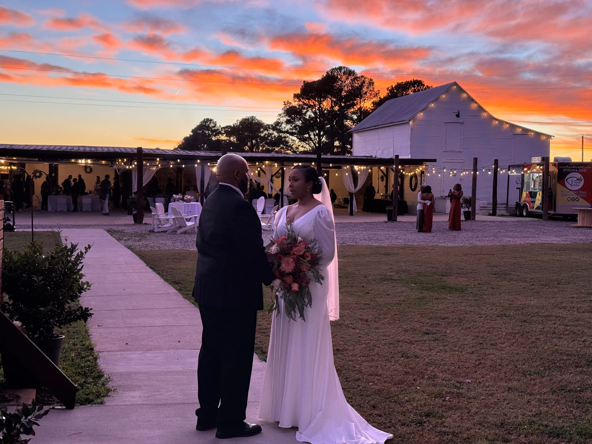 A bride holding a bouquet stands with her groom on a pathway as a sunset illuminates the white barn and string-lit pavilion at Scarborough House.