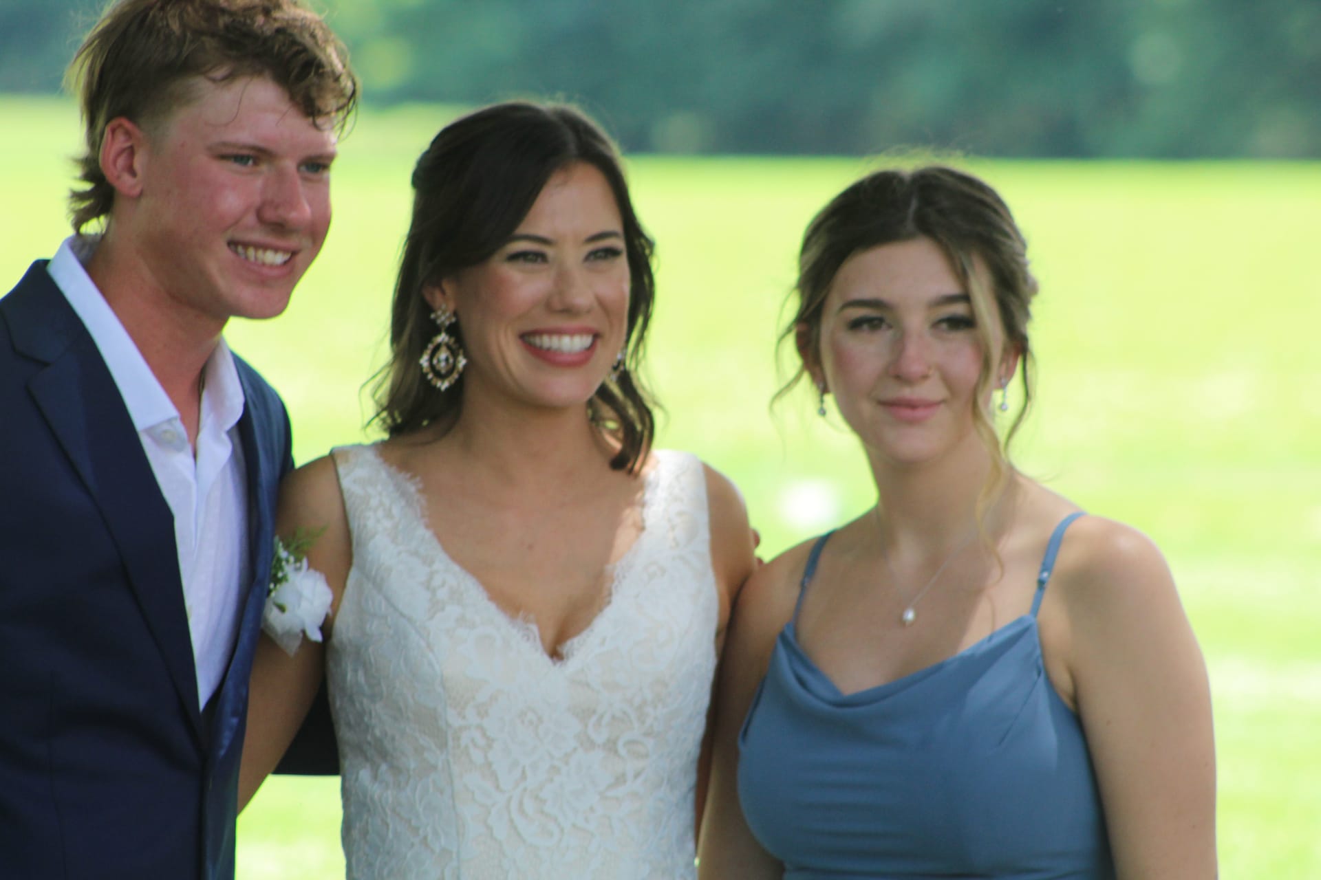 A smiling bride in a white lace gown stands between a young man in a navy suit and a bridesmaid in a dusty-blue dress against a sunlit green field at Scarborough House.
