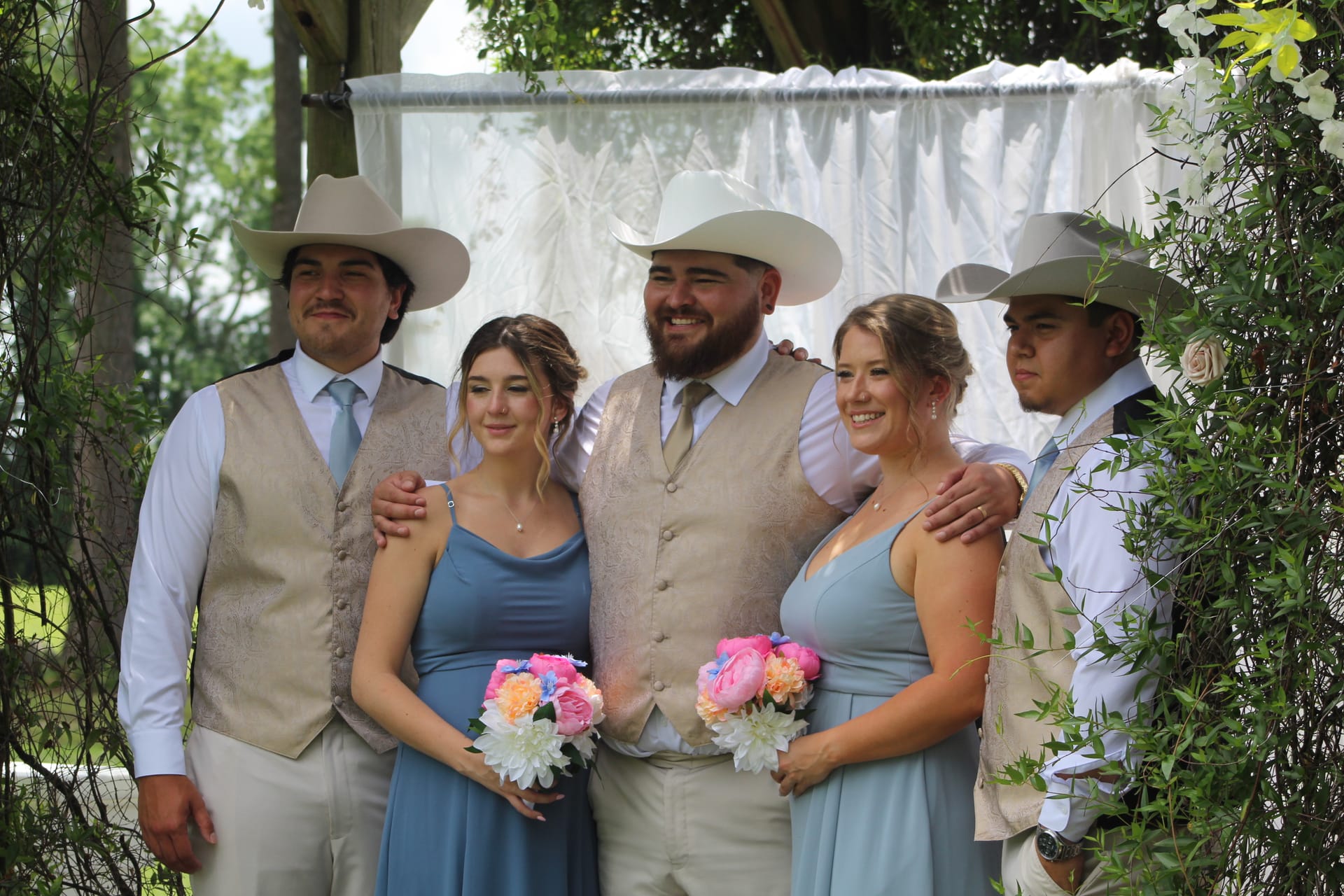 Groom in a tan vest and white cowboy hat stands with two bridesmaids in dusty blue dresses holding colorful bouquets and two groomsmen in matching vests and cowboy hats beneath a floral-draped ceremony arch at Scarborough House.