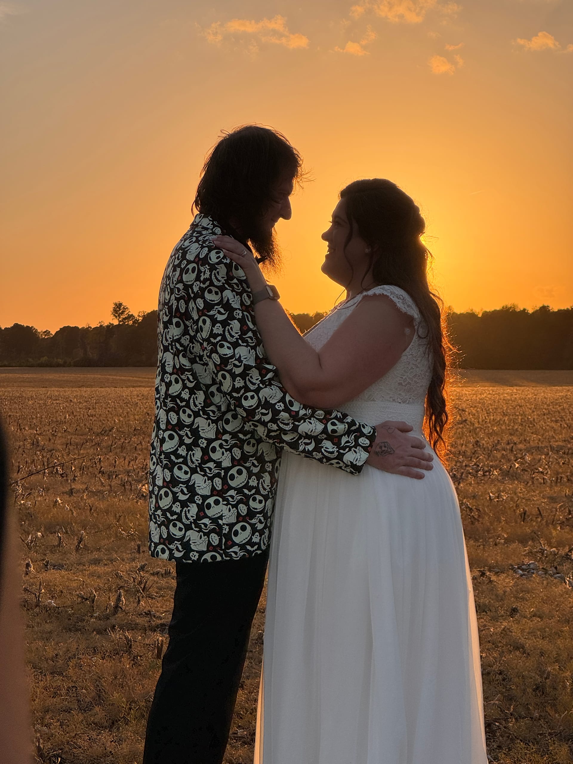 Bride in white lace gown and groom in a Nightmare Before Christmas printed shirt embrace facing each other against a vivid golden sunset over open farmland fields at Scarborough House.