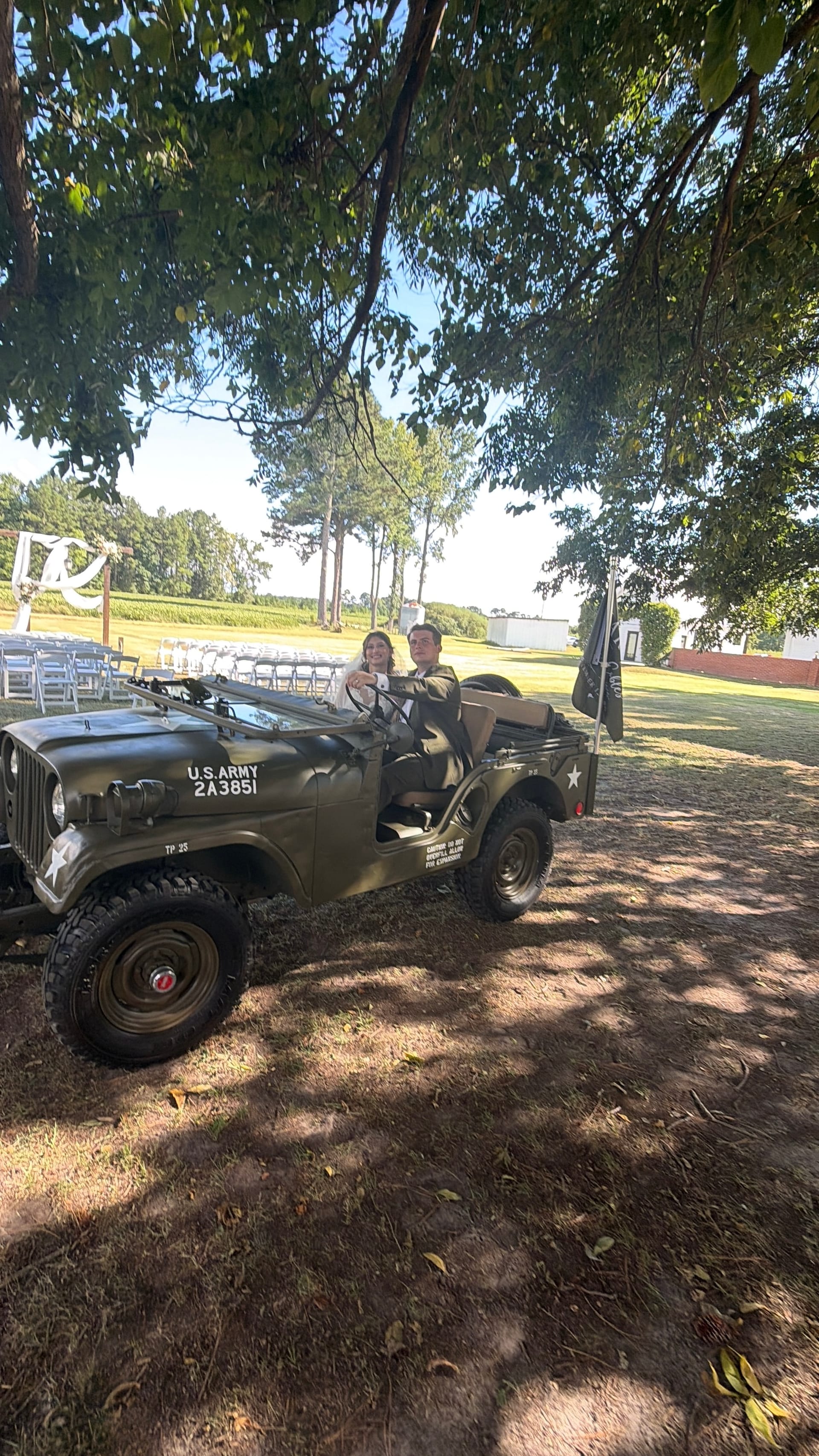 Bride and groom smiling together in a vintage olive green U.S. Army Jeep parked beneath oak trees on the Scarborough House estate grounds with a ceremony lawn visible in the background.