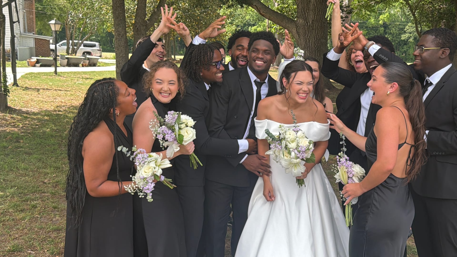 Bride in an off-shoulder white gown and groom in a dark suit laugh joyfully surrounded by their cheering wedding party dressed in black beneath sweeping oak trees.
