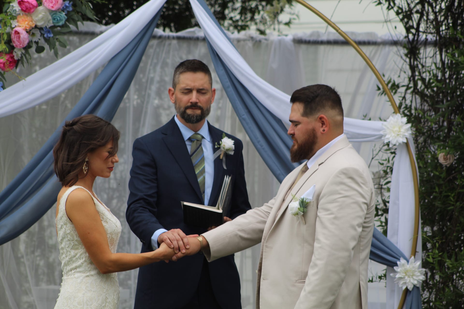 Bride and groom hold hands during wedding vow exchange before an officiant beneath a blue draped arch adorned with colorful florals at Scarborough House.