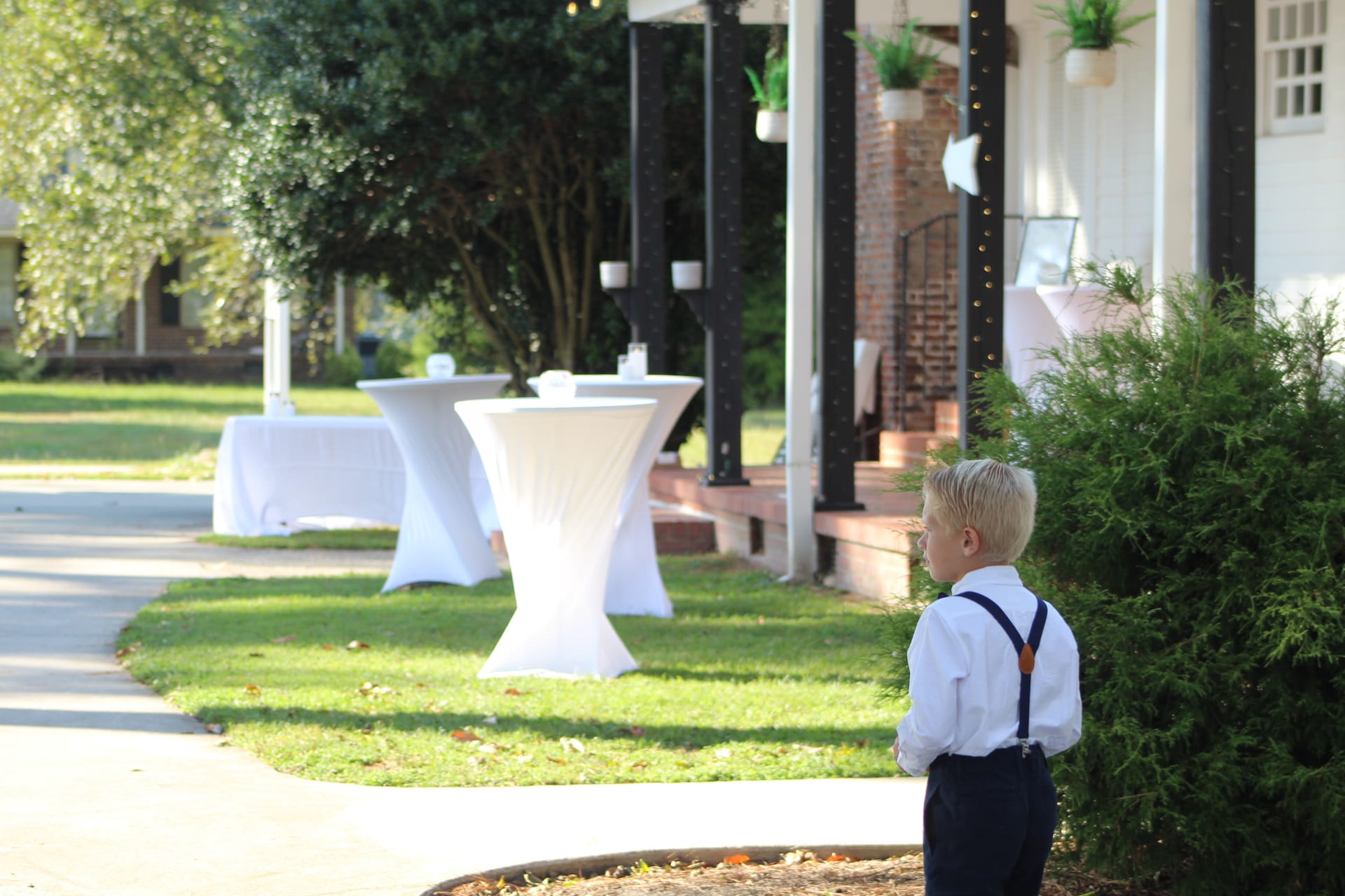 A young boy dressed in a white shirt and navy suspenders stands on the manicured lawn of Scarborough House looking toward white cocktail tables set up for an outdoor wedding reception.