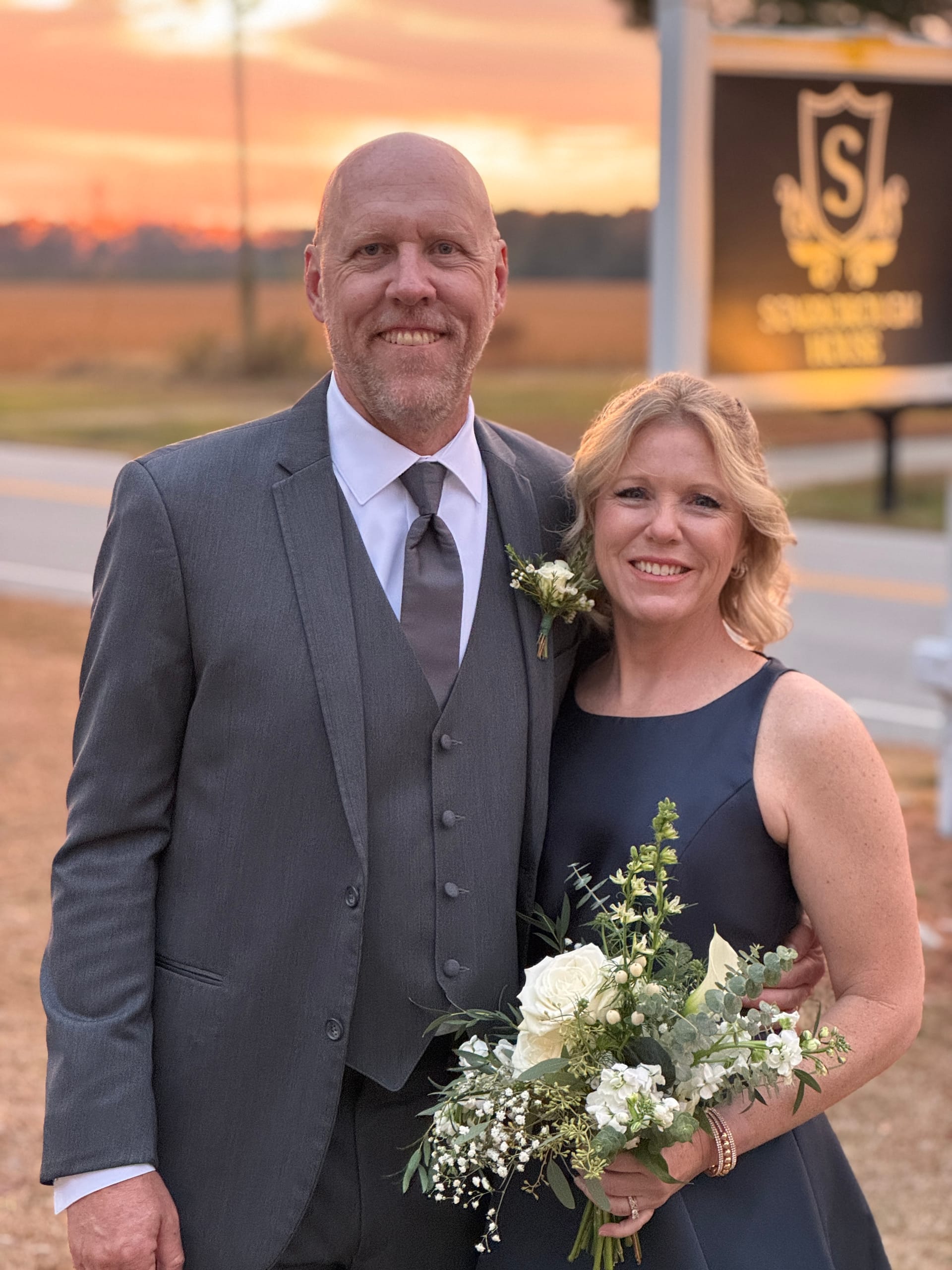 A smiling couple dressed in formal attire pose together at golden hour in front of the Scarborough House marquee sign, the woman holding a lush white and greenery bouquet against a vivid sunset sky.